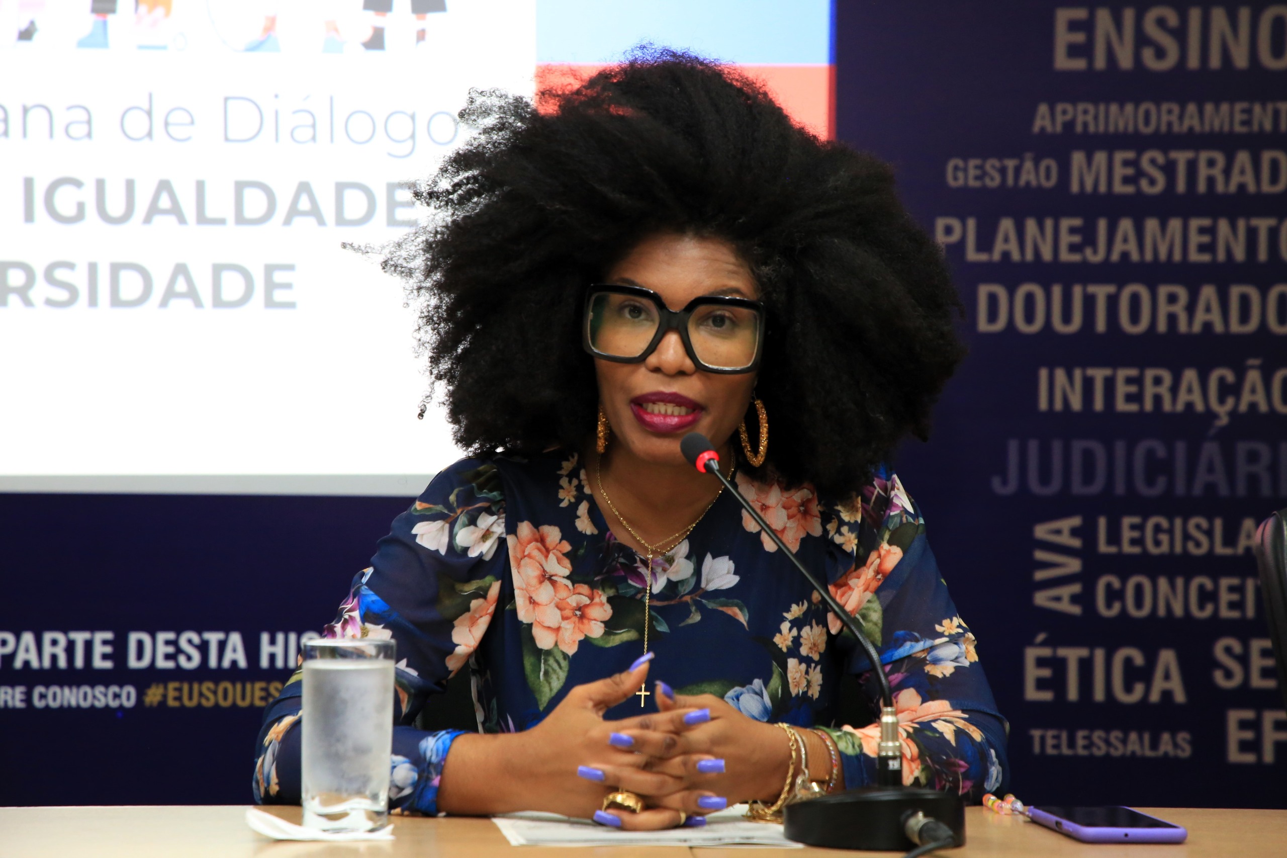 A foto mostra a juíza Renata, uma mulher negra, sentada à mesa, falando ao microfone. Ela usa óculos grandes, blusa floral e tem cabelo volumoso. Há um copo d’água à frente e um painel com textos ao fundo.
