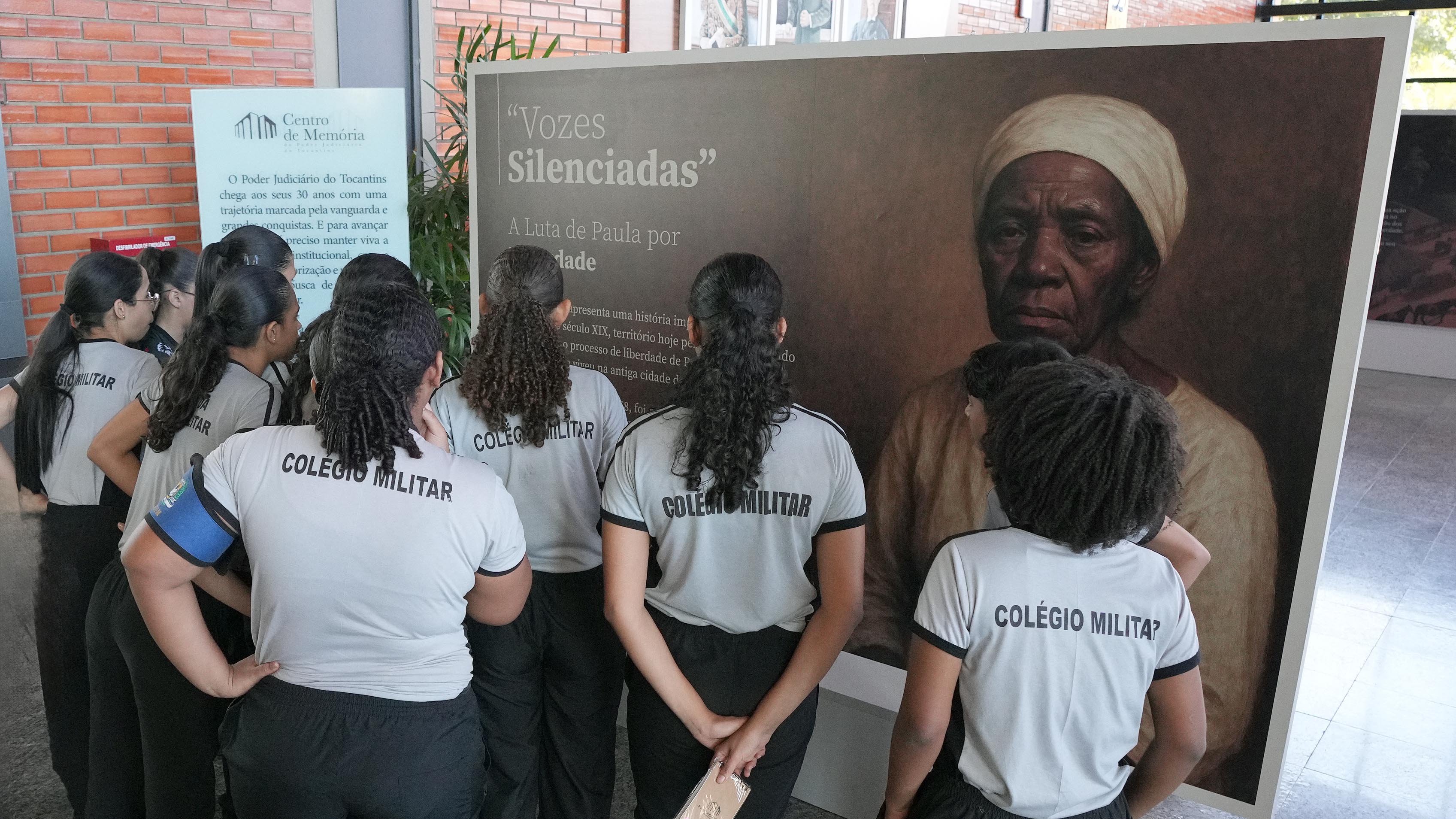 Um grupo de dez alunos estão na frente de um painel da mostra com o nome exposição vozes silenciadas. O quadro tem a foto de uma mulher negra. 