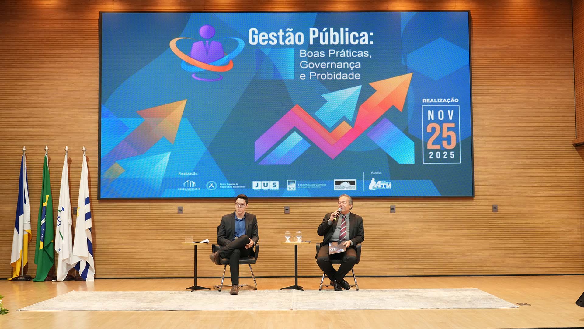A foto mostra o auditório do Tribunal de Justiça do Tocantins durante o Seminário Gestão Pública: Boas Práticas, Governança e Probidade. No centro do palco, dois homens estão sentados em poltronas pretas, cada um com uma mesa lateral de apoio com copos de água.  À esquerda, está João Paulo Landim Macedo, usando terno escuro, camisa azul e óculos, segurando um bloco de anotações sobre o colo. À direita, o juiz auxiliar da Presidência do TJTO, Ariostenes Guimarães Vieira, também de terno escuro, gravata bordô e microfone na mão, enquanto fala ao público.  Ao fundo, um grande telão ocupa toda a parede com o layout visual do evento, em tons de azul, roxo e laranja, com setas apontando para cima. No topo, lê-se o título “Gestão Pública: Boas Práticas, Governança e Probidade”