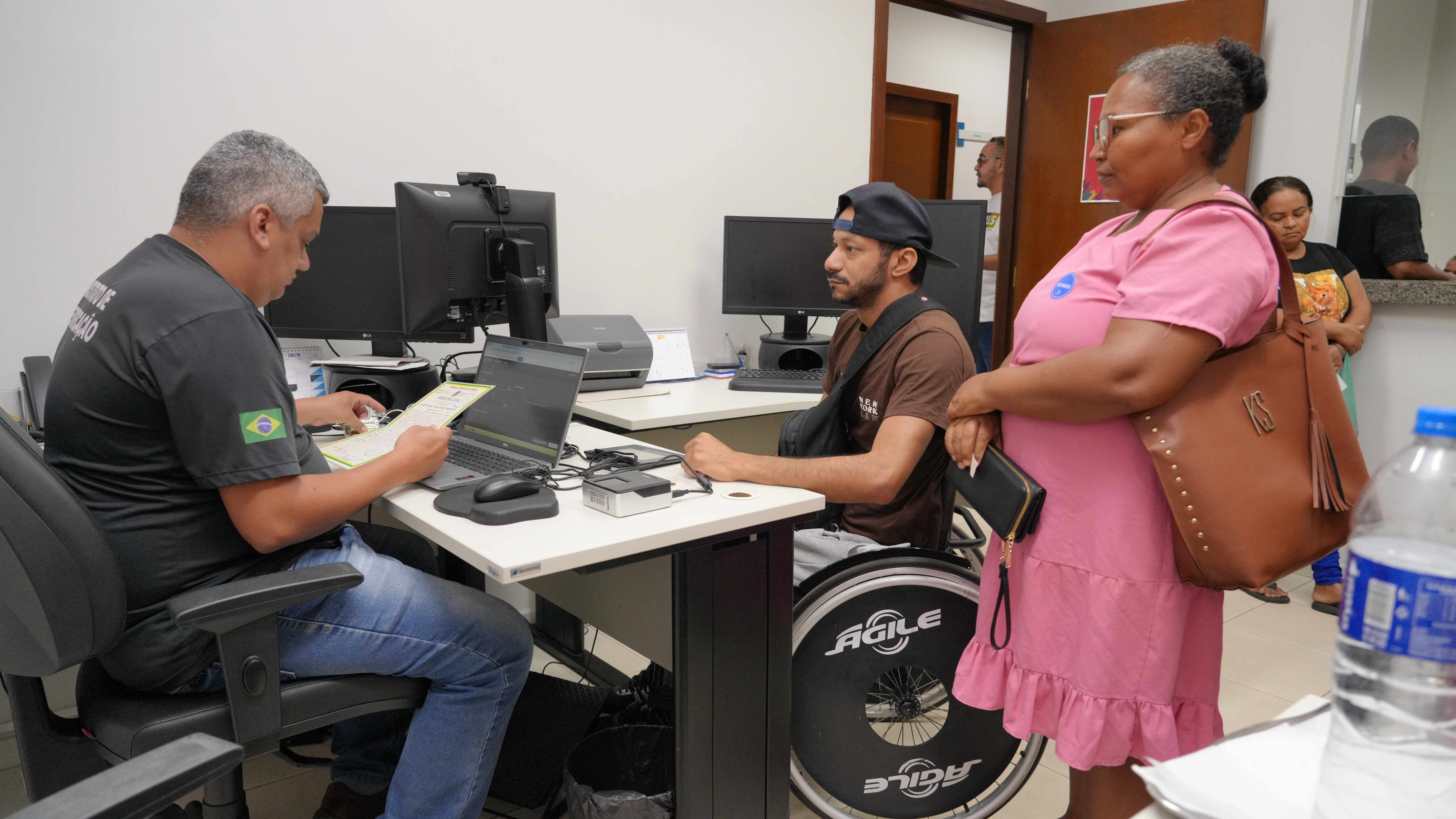 A foto mostra um atendente analisando um documento diante de um homem em cadeira de rodas. Ao lado dele, uma mulher em pé aguarda. Ao fundo, outras pessoas também esperam.