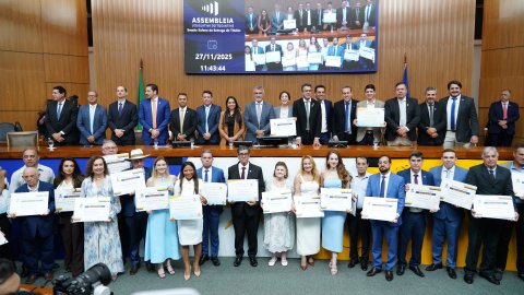 A foto registra o momento oficial da entrega dos títulos na Assembleia Legislativa do Tocantins. No centro do plenário, dezenas de homenageados posam em pé, lado a lado, segurando seus certificados. Atrás deles, parlamentares e autoridades ocupam a mesa diretora, formando a segunda fileira do registro. Um grande painel eletrônico exibe a data — 27/11/2025 — e imagens do próprio evento.
