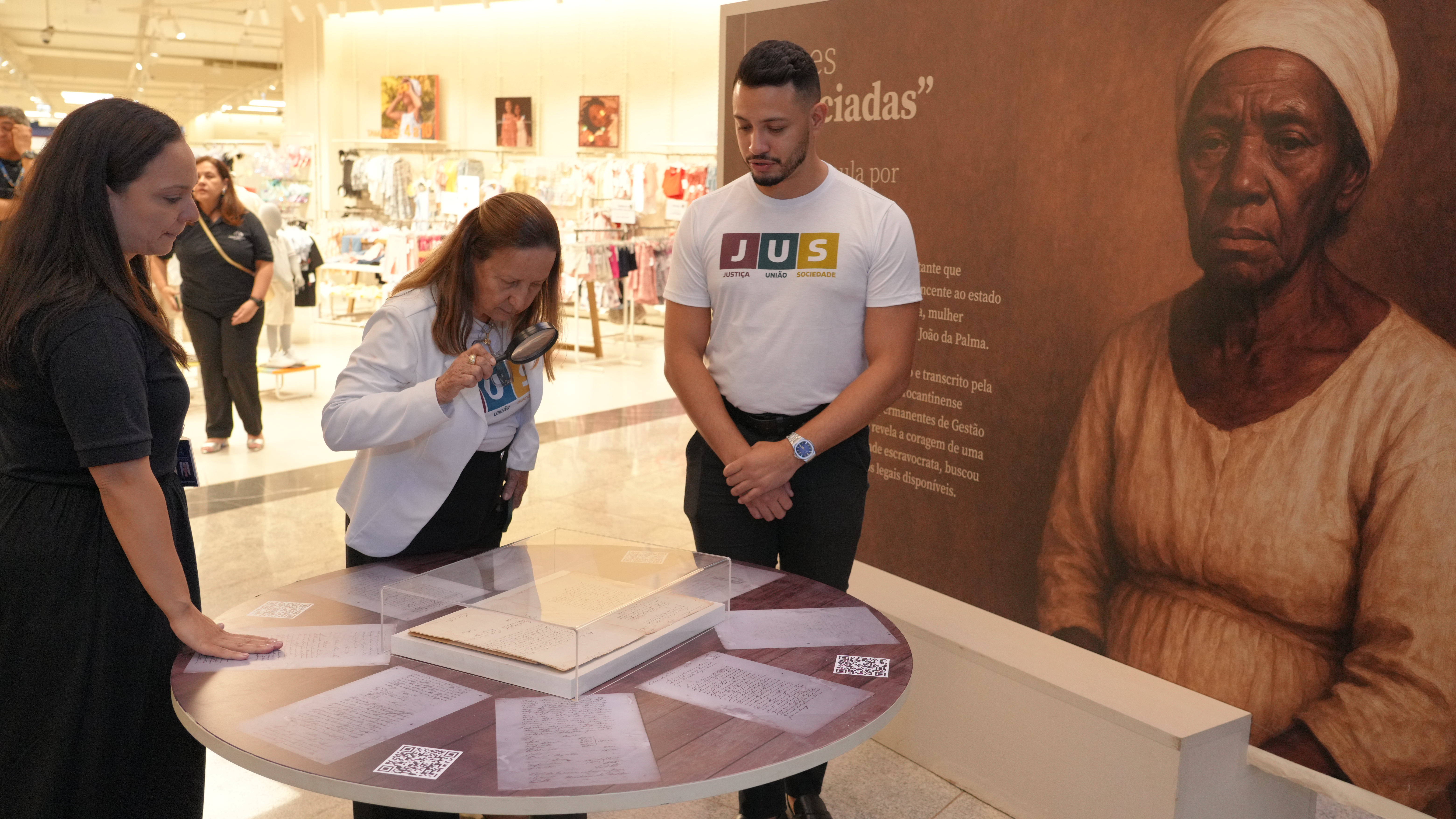 A presidente do TJTO, desembargadora Maysa Vendramini,  observa documentos expostos sobre uma mesa; ela está ao centro e usa uma lupa, observada por uma mulher (esq) e um homem (dir). À direita, há um painel com a imagem de uma mulher negra. O cenário é um shopping, com vitrines ao fundo.
