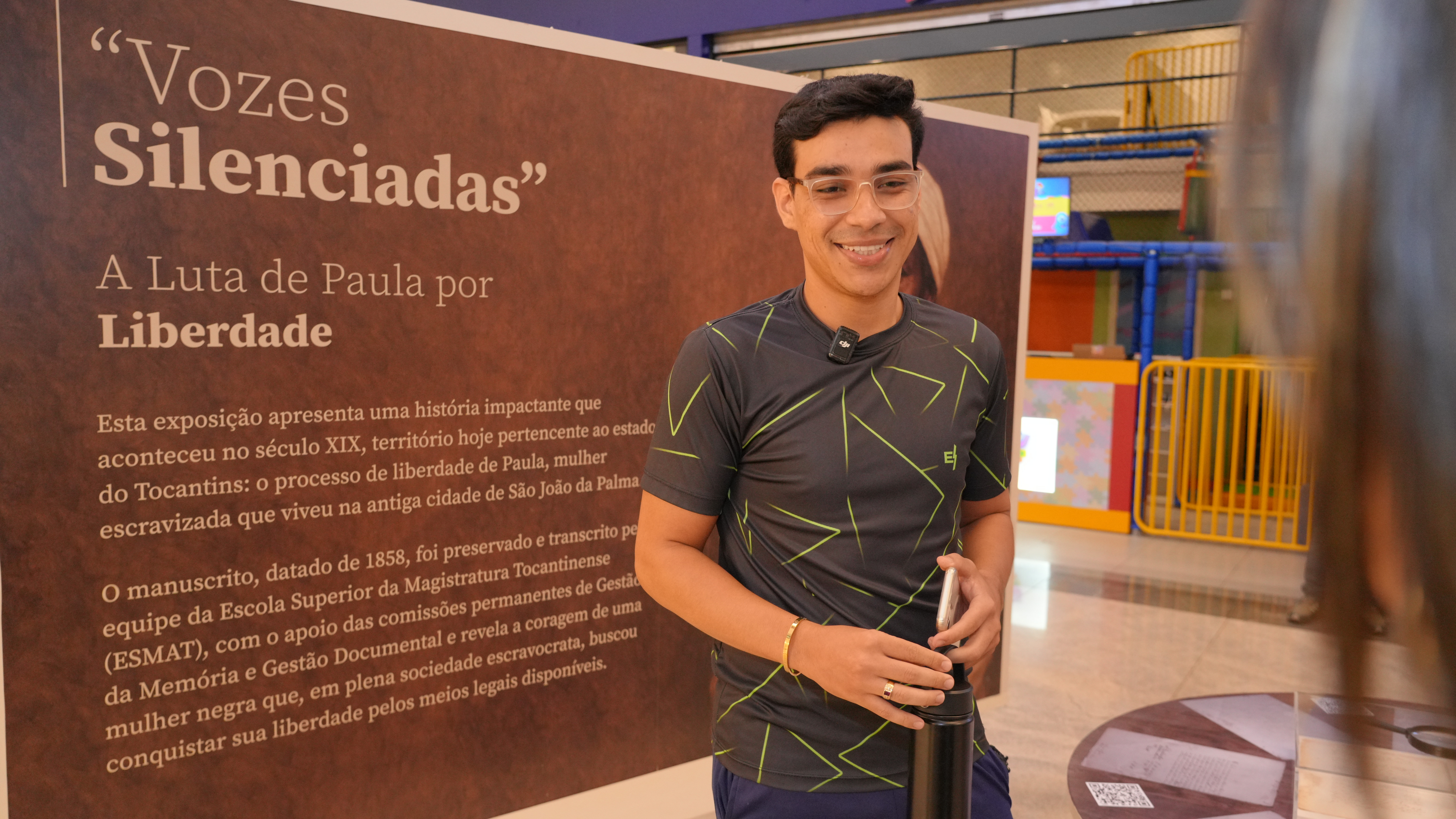 Um homem sorri enquanto segura uma garrafa e um celular, posicionado ao lado do painel da exposição “Vozes Silenciadas: A Luta de Paula por Liberdade”. O texto do painel aparece parcialmente ao fundo. O ambiente é interno, com estruturas coloridas de área infantil visíveis atrás.
