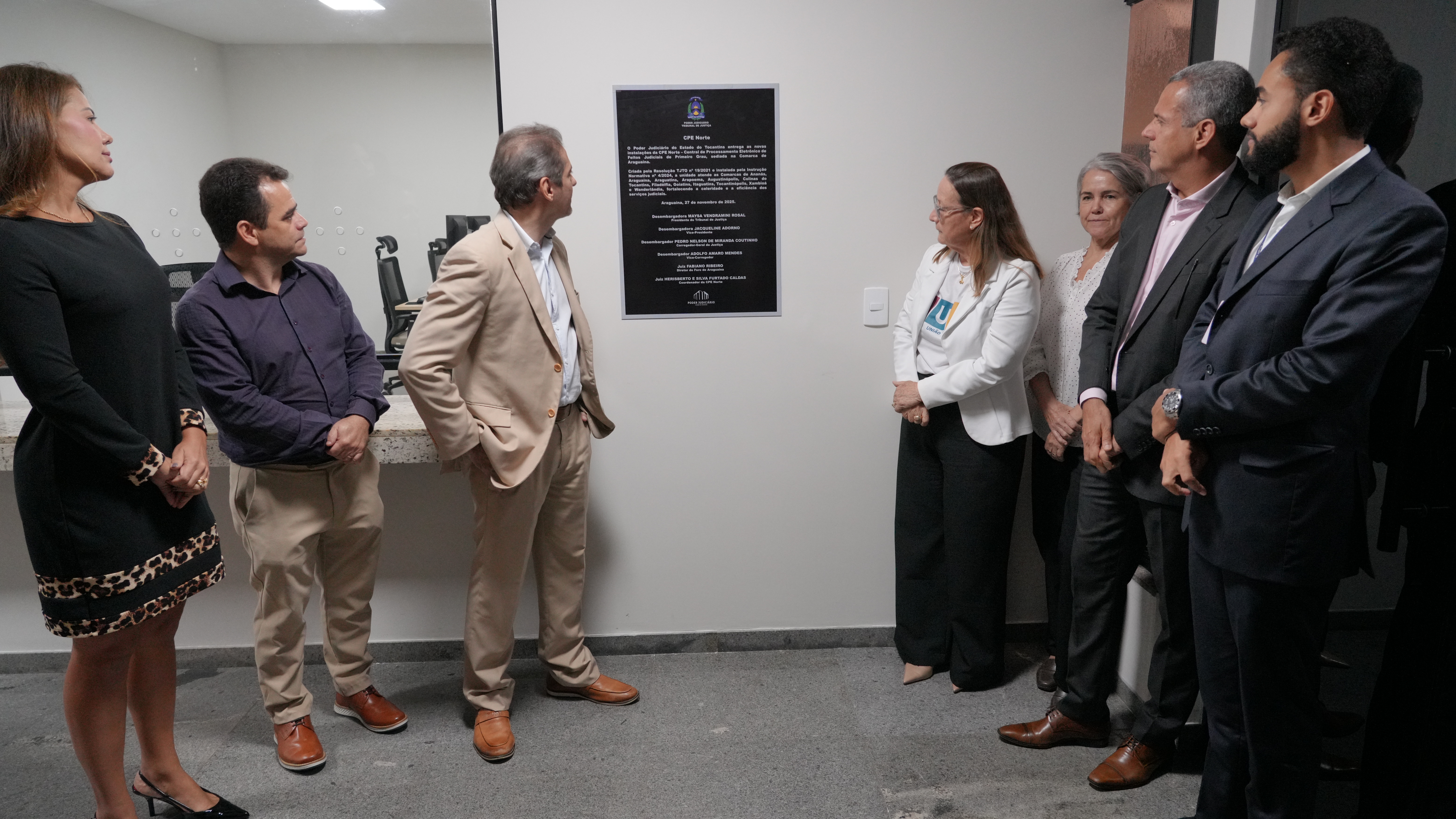 Um grupo de sete pessoas participa de um momento solene de inauguração ou descerramento de placa em um ambiente institucional. Elas estão posicionadas em semicírculo, voltadas para uma placa preta fixada na parede, que contém texto formal e o brasão do Tribunal de Justiça no topo. O ambiente é interno, com paredes claras, piso de granito e bancada ao fundo, sugerindo que o espaço pertence a uma unidade administrativa recém-inaugurada ou revitalizada. A cena transmite formalidade, respeito e importância institucional.