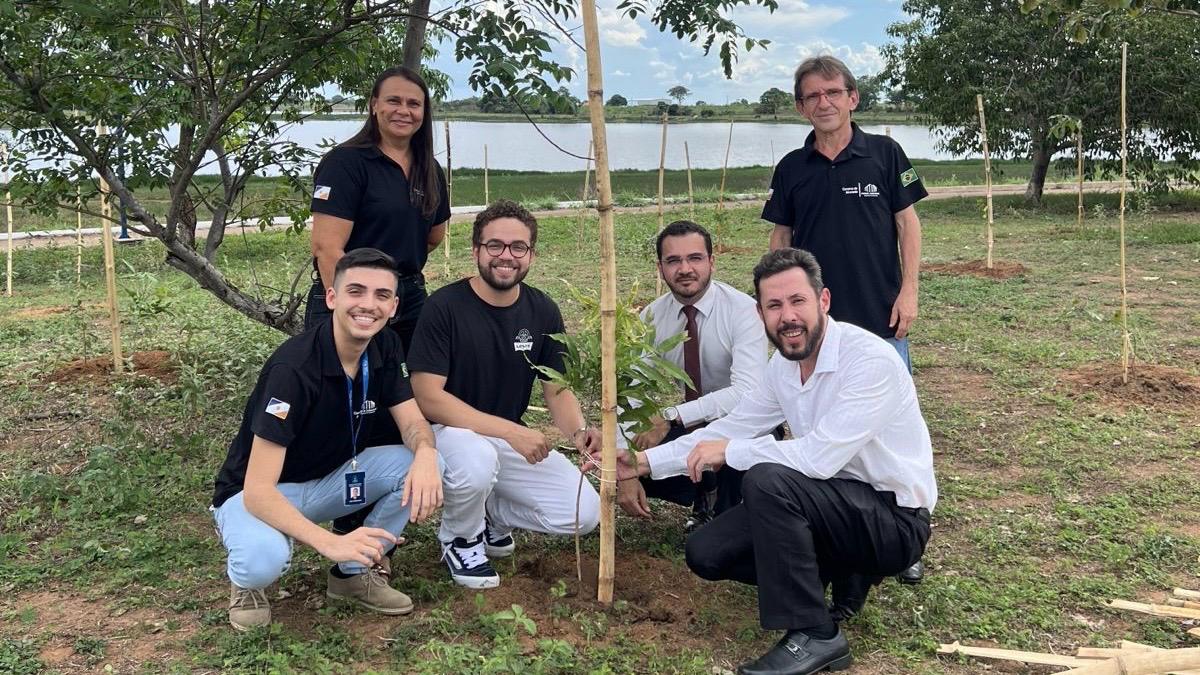 Seis pessoas posam em um ambiente aberto, durante ação de plantio de mudas às margens de uma lagoa. Em primeiro plano, quatro homens estão agachados ao redor de uma muda recém-plantada, segurando o pequeno caule e demonstrando participação ativa na atividade. Eles usam roupas sociais ou camisetas pretas com identificação institucional.  Ao fundo, uma mulher e um homem — ambos de camiseta preta — permanecem em pé, sorrindo e acompanhando o registro.