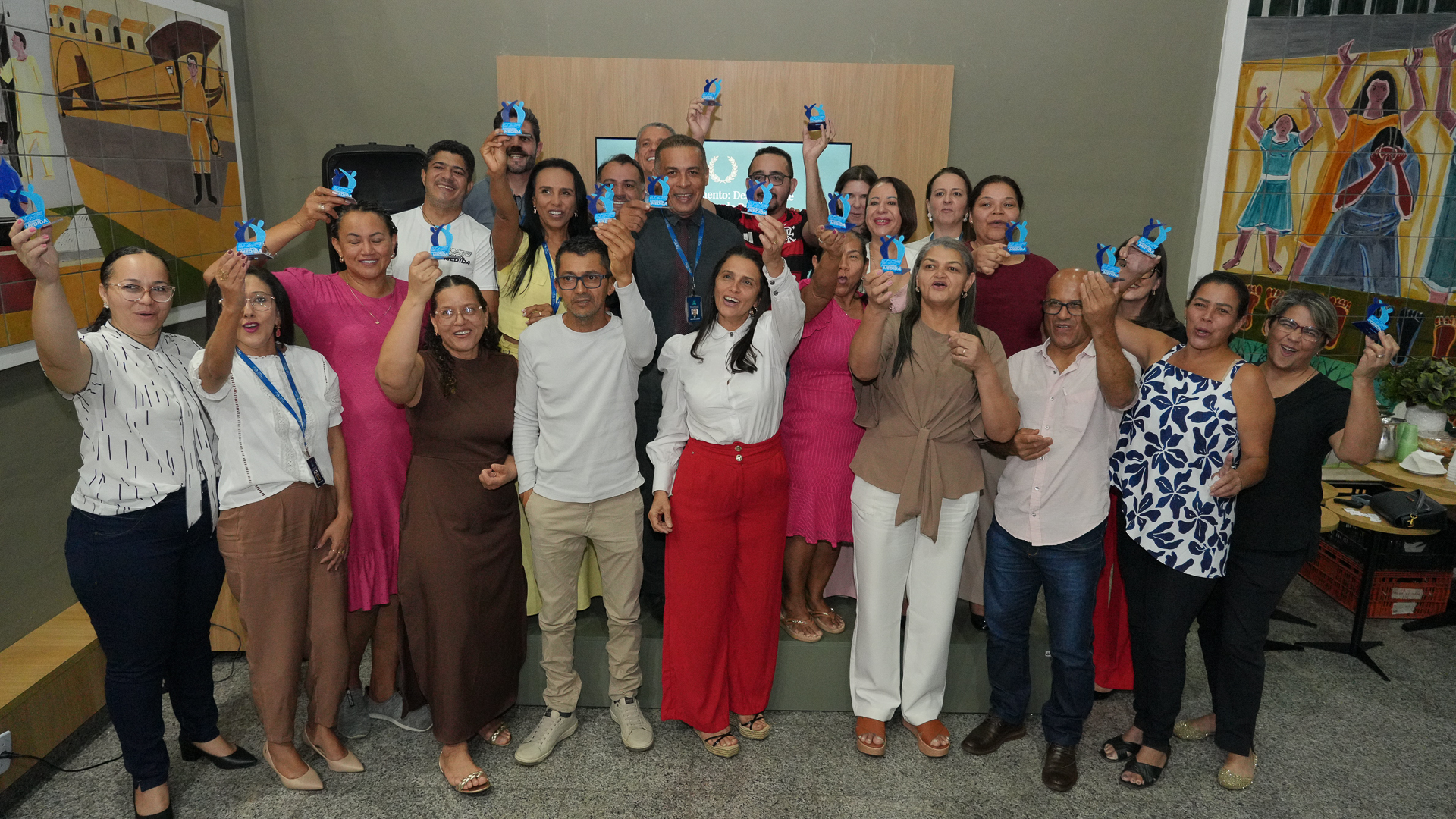 A foto mostra um grupo grande de pessoas reunidas, posando para a câmera e levantando pequenos troféus azuis. Todos sorriem e celebram juntos em um ambiente interno, com murais coloridos nas paredes ao fundo. O clima é de confraternização e reconhecimento coletivo.