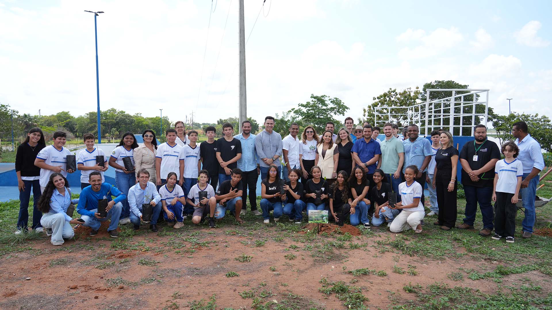 A foto mostra um grande grupo reunido em área aberta, durante uma ação ambiental de plantio de mudas. Ao centro e nas laterais, há adultos e jovens — muitos deles estudantes com uniformes escolares brancos e detalhes azuis — todos posicionados para a foto. Várias pessoas na fileira da frente estão agachadas segurando mudas em pequenos sacos pretos.