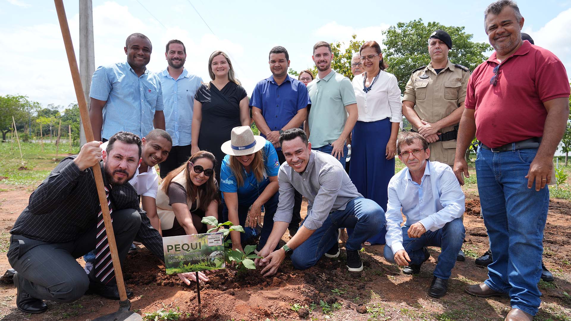 A foto registra um momento de plantio de uma muda de pequi. Em primeiro plano, um grupo de pessoas está agachado ao redor da pequena planta, tocando a terra e sorrindo para a câmera. Ao lado da muda há uma placa identificando a espécie: “PEQUI (Caryocar brasiliense)”.  As pessoas envolvidas representam diferentes instituições e perfis profissionais — há homens e mulheres, alguns com roupas sociais, outros com uniforme azul, além de um policial militar em trajes oficiais. À esquerda, um dos participantes segura uma enxada, reforçando o caráter simbólico e colaborativo da ação.