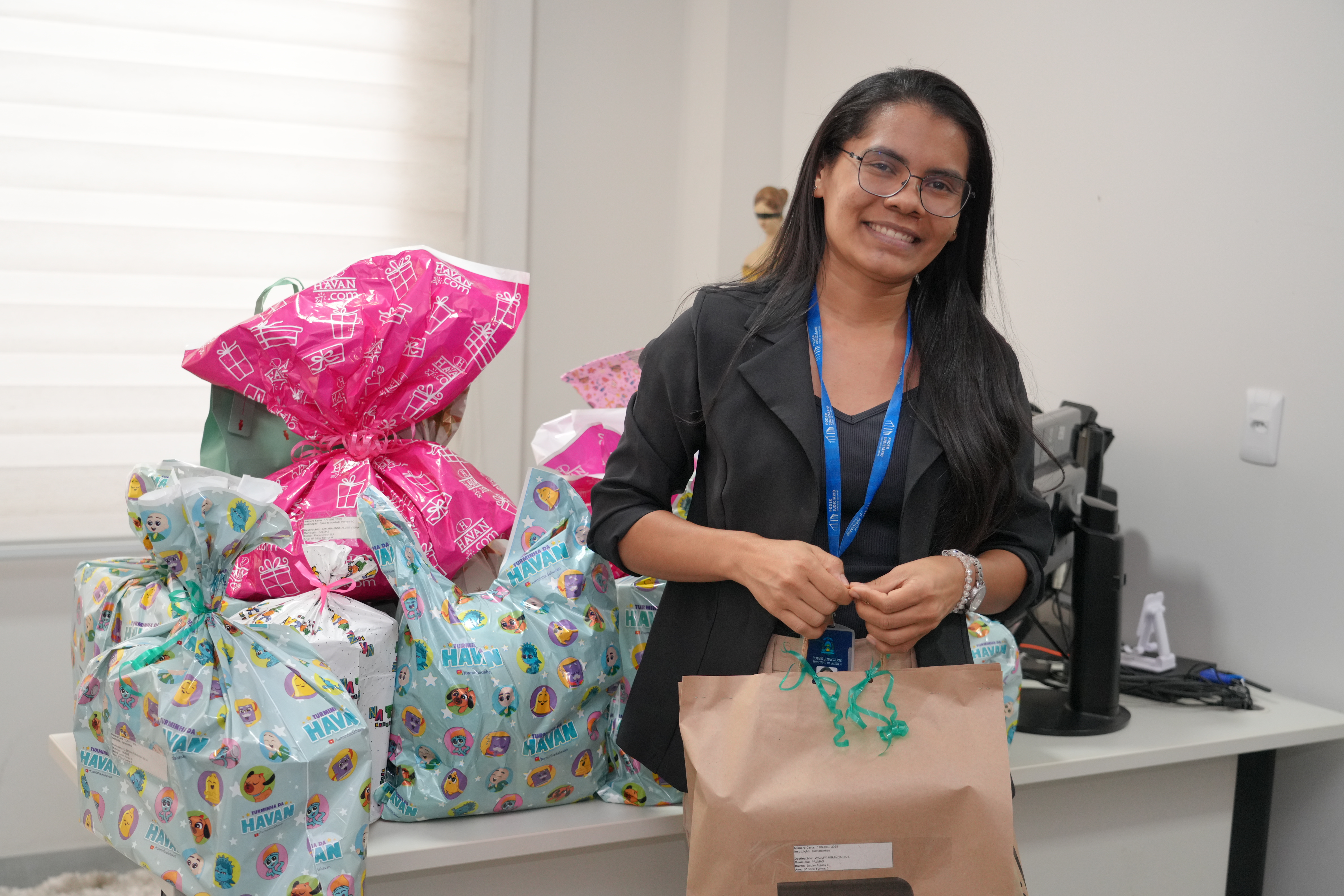 A foto mostra uma mulher de crachá azul sorrindo, em pé ao lado de uma mesa cheia de presentes embrulhados em papéis coloridos. Ela segura uma sacola de papel pardo com um laço verde.
