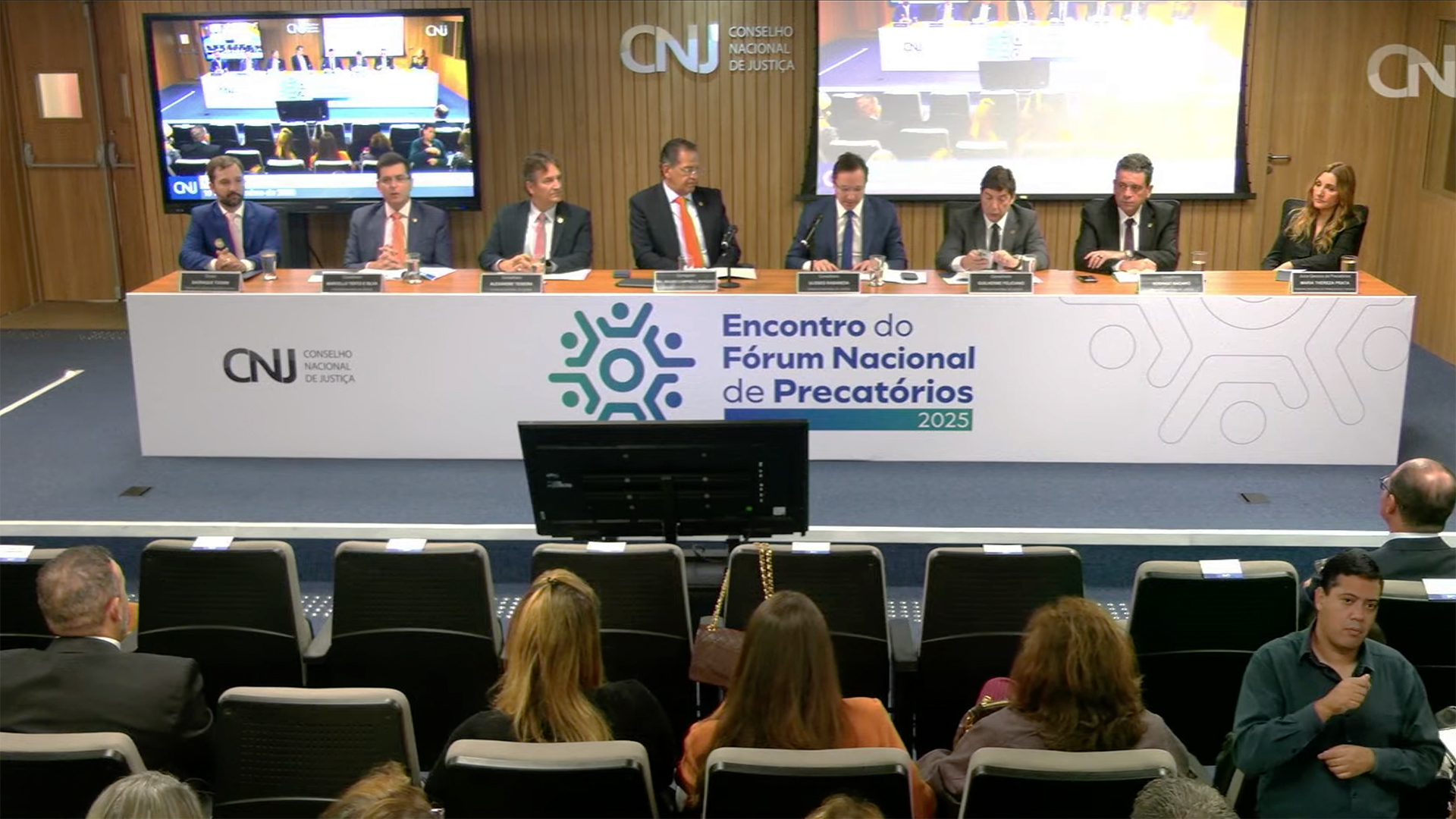 A imagem mostra uma mesa com oito autoridades reunidas no auditório do Conselho Nacional de Justiça (CNJ), durante o “Encontro do Fórum Nacional de Precatórios 2025”. Elas estão posicionadas à frente de uma mesa identificada com a marca do evento, enquanto um telão ao fundo exibe a transmissão. Na parte inferior da imagem, parte da plateia acompanha a programação.