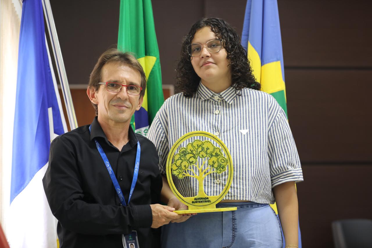 Duas pessoas posam segurando um troféu com o símbolo de uma árvore e a inscrição “Alvorada Sustentável”. Ao fundo, bandeiras oficiais reforçam o contexto de reconhecimento ligado à sustentabilidade e responsabilidade ambiental.