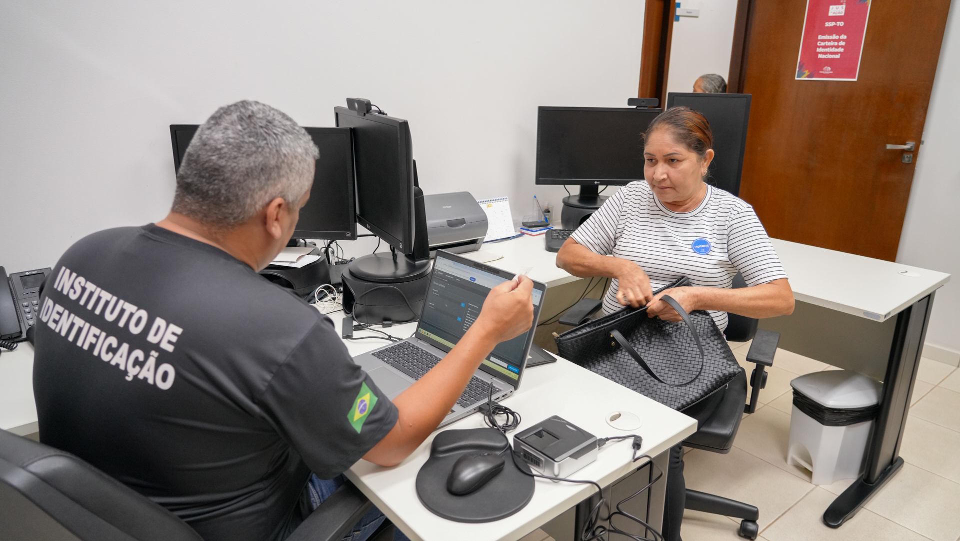 Atendimento para emissão de documento de identidade: um servidor do Instituto de Identificação realiza o procedimento em computador, enquanto uma cidadã organiza seus pertences e recebe orientações.