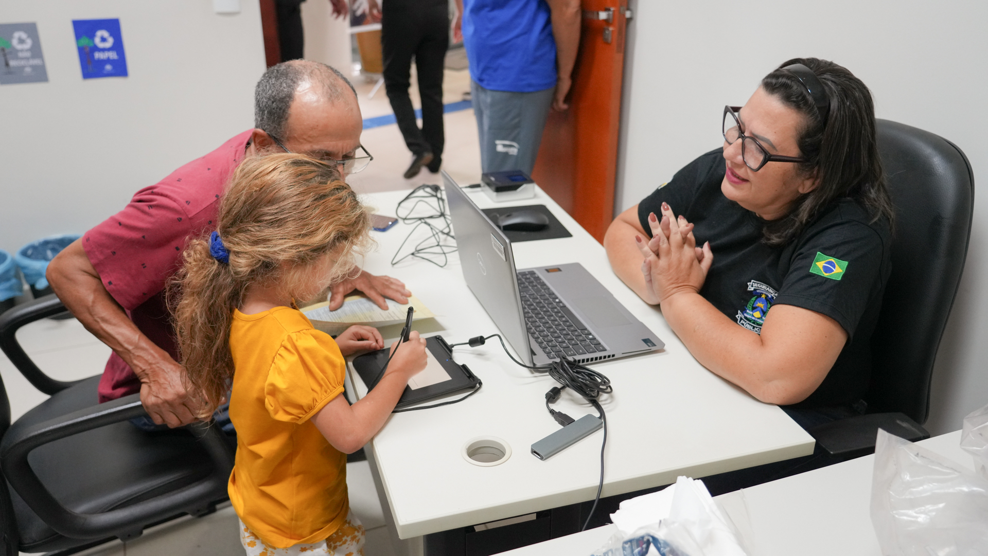 Atendimento institucional em mesa de serviço: uma servidora orienta um homem e uma criança durante procedimento de cadastro, com uso de computador e mesa digital para assinatura. O responsável acompanha atentamente enquanto a criança realiza o registro, em ambiente organizado e voltado ao atendimento ao público.