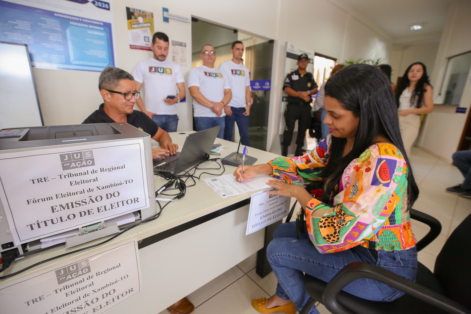 Atendimento do Tribunal Regional Eleitoral durante ação do JUS em Ação: uma mulher preenche formulário para emissão do título de eleitor em balcão identificado do TRE, enquanto um servidor realiza o cadastro em computador. Ao fundo, servidores do Judiciário acompanham a atividade, além de um agente de segurança, em ambiente institucional organizado e sinalizado.