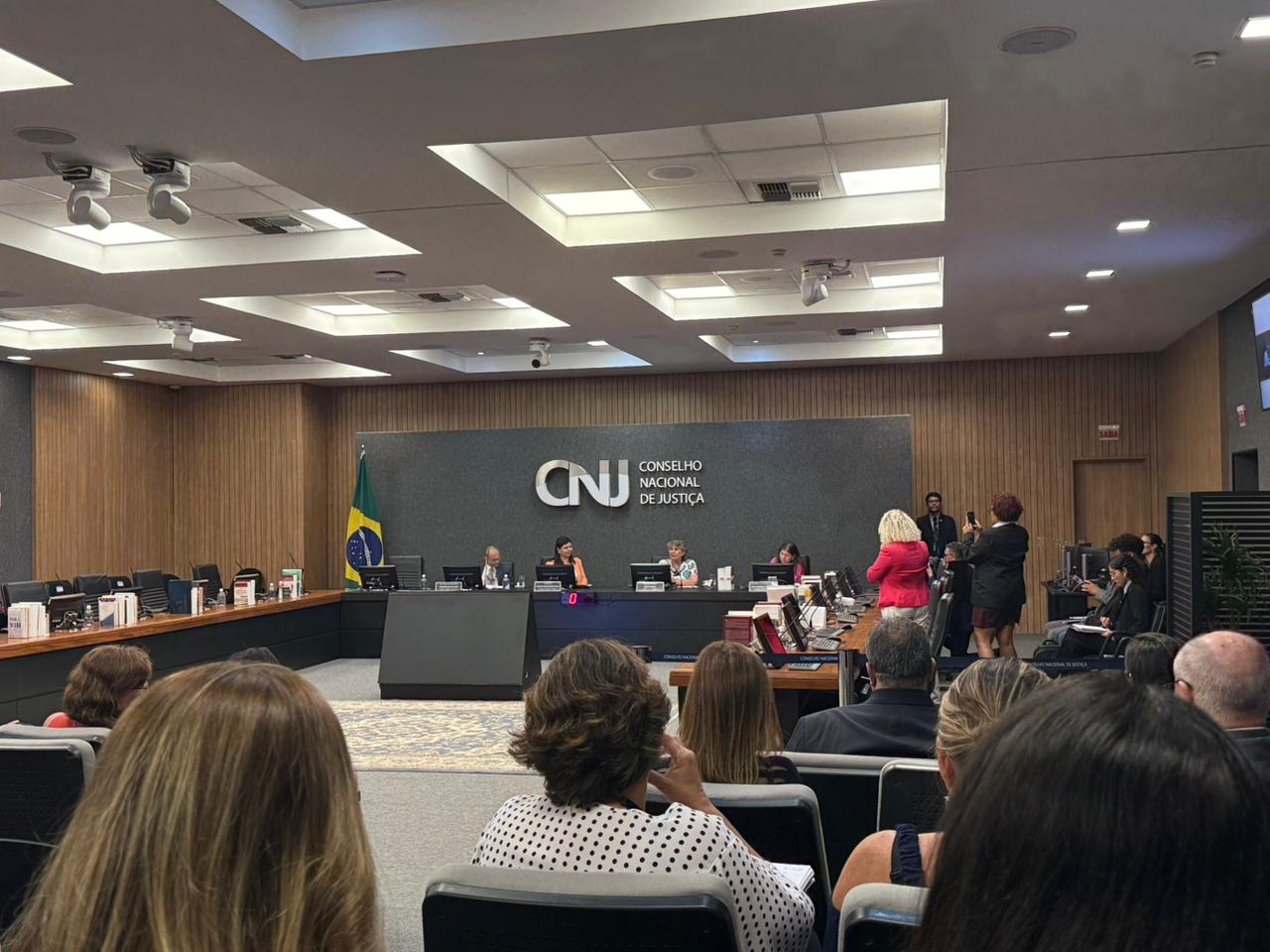 A imagem mostra uma sessão realizada no auditório do Conselho Nacional de Justiça (CNJ), em Brasília. No centro do espaço, vê-se a mesa diretora, com autoridades sentadas à frente do plenário, tendo ao fundo o painel com a logomarca do CNJ – Conselho Nacional de Justiça e a bandeira do Brasil.  O ambiente é amplo, moderno e bem iluminado, com iluminação embutida no teto e revestimento em madeira nas paredes laterais. À frente, integrantes da mesa conduzem os trabalhos, enquanto servidores, magistrados e convidados acompanham a sessão sentados no plenário. Algumas pessoas estão em pé, registrando o momento com celulares, o que indica a relevância institucional do evento.
