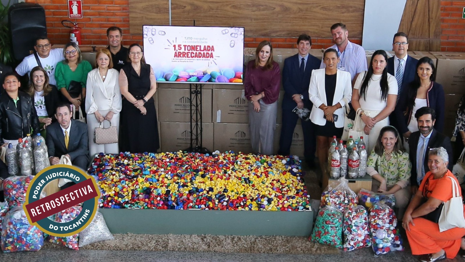 Grupo de pessoas participa de ação solidária e ambiental, posando ao lado de grande volume de tampinhas plásticas arrecadadas que foram doadas para o hospital do Amor. O painel ao fundo destaca o resultado da campanha, com  uma tonelada e meia de tampinhas arrecadadas. 