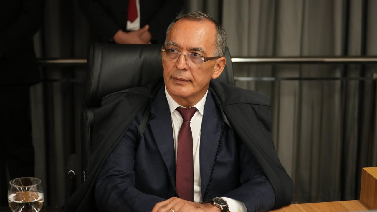 Desembargador Gil, sentado em cadeira institucional, atrás de mesa de trabalho, em ambiente formal. Usa traje social escuro, gravata e toga sobre os ombros. Mantém postura ereta, com as mãos apoiadas à frente, e olha para o lado. Sobre a mesa, há um microfone e um copo com água. Ao fundo, cortinas em tom neutro e pessoas em pé compõem o cenário solene.