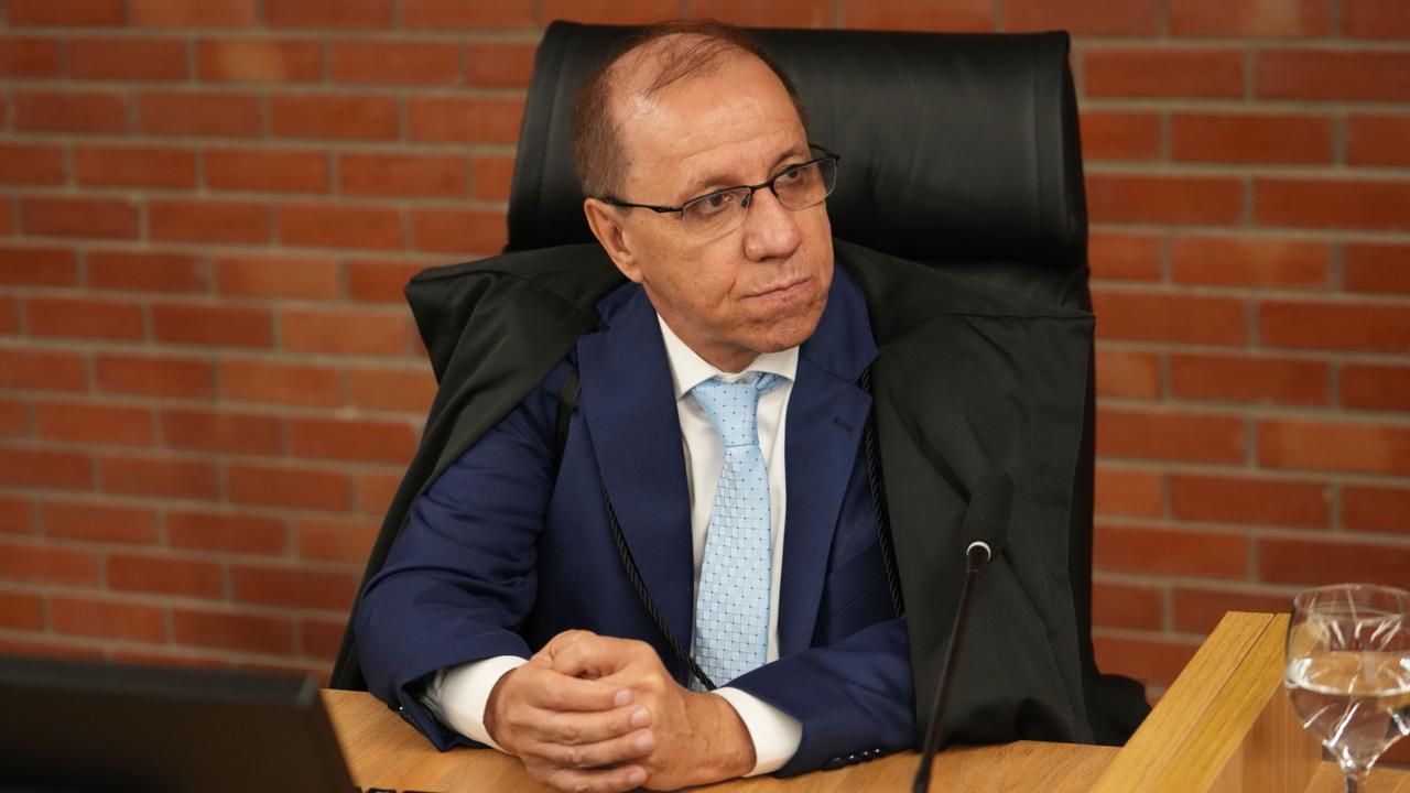 Desembargador Luiz Zilmar, sentado em cadeira institucional, atrás de mesa de trabalho, em ambiente formal. Usa óculos, traje social azul com gravata clara e toga sobre os ombros. Mantém as mãos entrelaçadas sobre a mesa e olha para o lado. À frente, há um microfone e um copo com água. O fundo apresenta parede de tijolos aparentes.