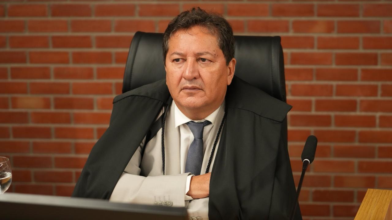 Desembargador Nelson Coelho, sentado em cadeira institucional, atrás de mesa de trabalho, em ambiente formal. Usa traje social claro com gravata e toga sobre os ombros. Mantém postura ereta, com expressão séria, olhando para o lado. À direita, há um microfone, e ao fundo aparece parede de tijolos aparentes.
