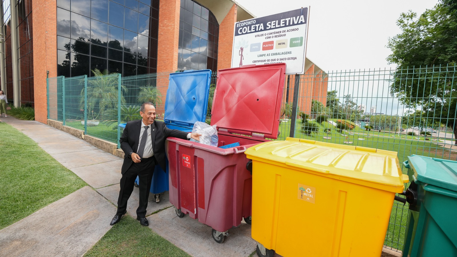 A imagem mostra um homem de terno descartando resíduos em contêineres de coleta seletiva coloridos (azul, vermelho, amarelo e verde), posicionados em área externa próxima a um prédio institucional, com placa informativa sobre reciclagem ao fundo.