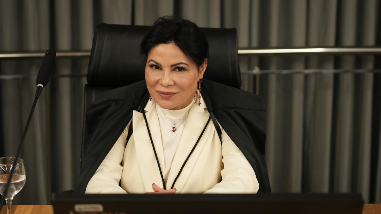 Desembargadora Edilene, sentada em cadeira institucional, atrás de mesa de trabalho, em ambiente formal. Usa traje claro, com toga preta sobre os ombros e acessórios discretos. Mantém postura ereta, com as mãos apoiadas à frente. Ao lado, há um microfone e um copo com água, com fundo de cortinas em tom neutro.