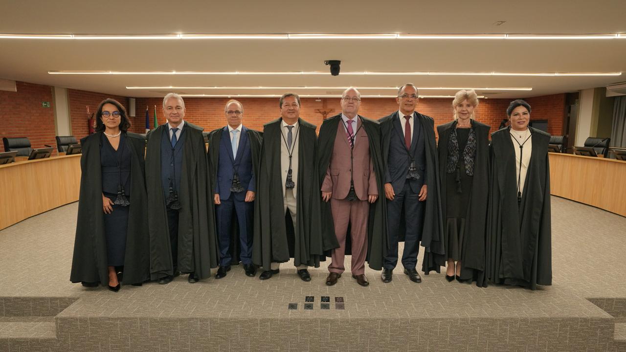 Os oito novos desembargadores do Tribunal de Justiça do Tocantins estão posicionados lado a lado, em pé, no plenário do TJTO, para registro oficial. Todos vestem trajes formais e togas, em ambiente solene. Ao fundo, aparecem as bancadas do plenário, com mesas, cadeiras e iluminação linear no teto, além de paredes em tijolos aparentes, reforçando o caráter institucional da cerimônia.