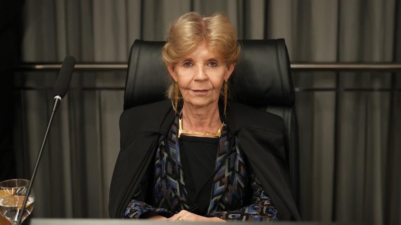 Desembargadora Silvana, mulher sentada em cadeira institucional, atrás de mesa de trabalho, em ambiente formal. Veste traje escuro com acessório dourado, mantém postura séria e mãos apoiadas à frente. Ao lado, há um microfone e um copo com água, com fundo de cortinas em tom neutro.