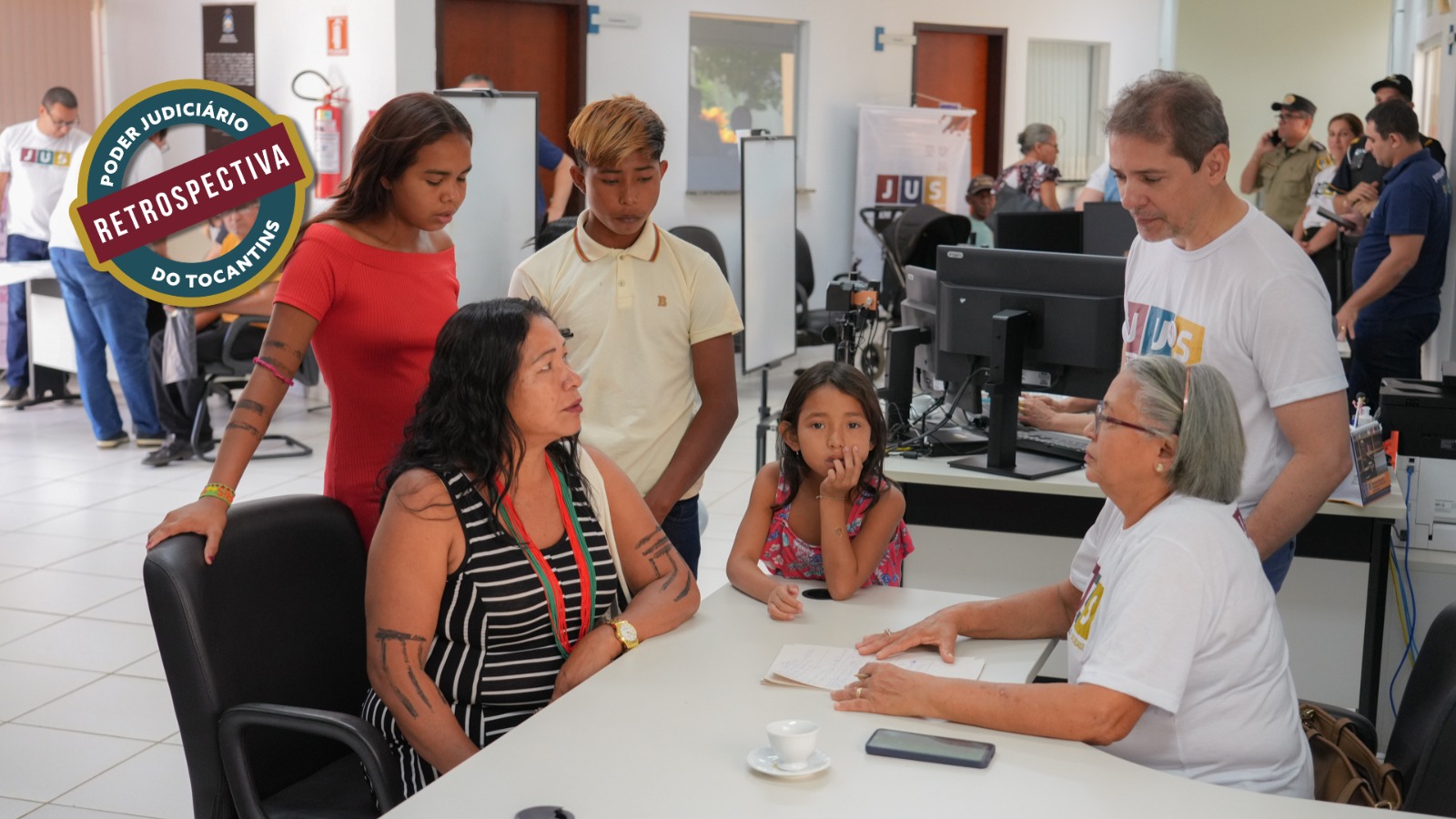 A foto mostra atendimento do Poder Judiciário do Tocantins a uma família, em mesa equipada com computador e documentos. Uma servidora orienta os cidadãos, enquanto adultos e crianças acompanham o atendimento. Ao fundo, outros servidores realizam atendimentos simultâneos em ambiente institucional organizado.
