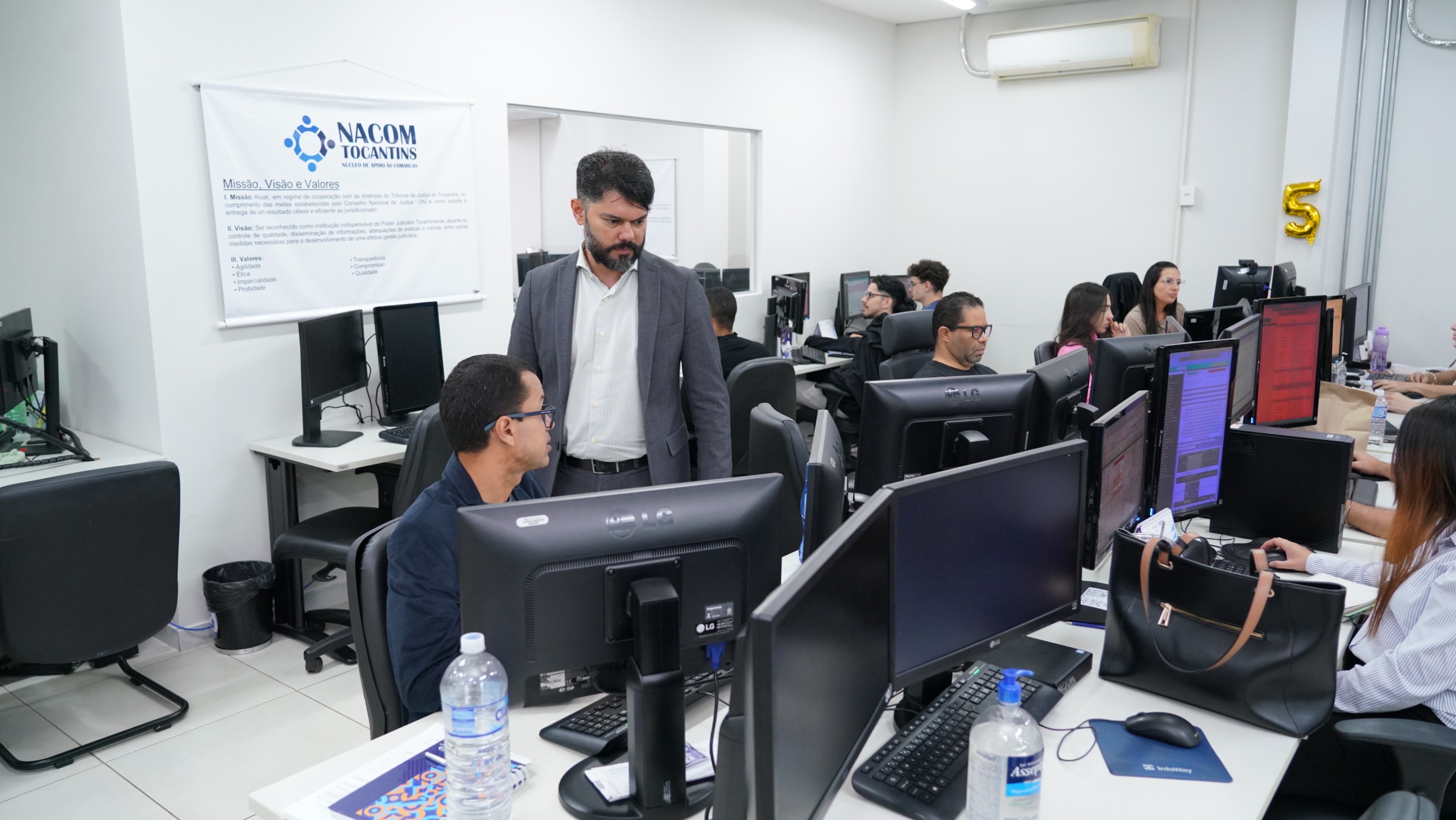 A imagem mostra o ambiente de trabalho do Núcleo de Apoio às Comarcas do Tribunal de Justiça do Tocantins (Nacom/TJTO), com servidores(as) atuando de forma integrada em estações de trabalho organizadas e equipadas com computadores.  Em destaque, um magistrado acompanha e orienta o trabalho de um servidor, evidenciando a proximidade da gestão com a equipe. Ao fundo, um banner institucional do Nacom apresenta a missão, visão e valores do núcleo, reforçando o compromisso com a eficiência, a organização e a qualidade da prestação