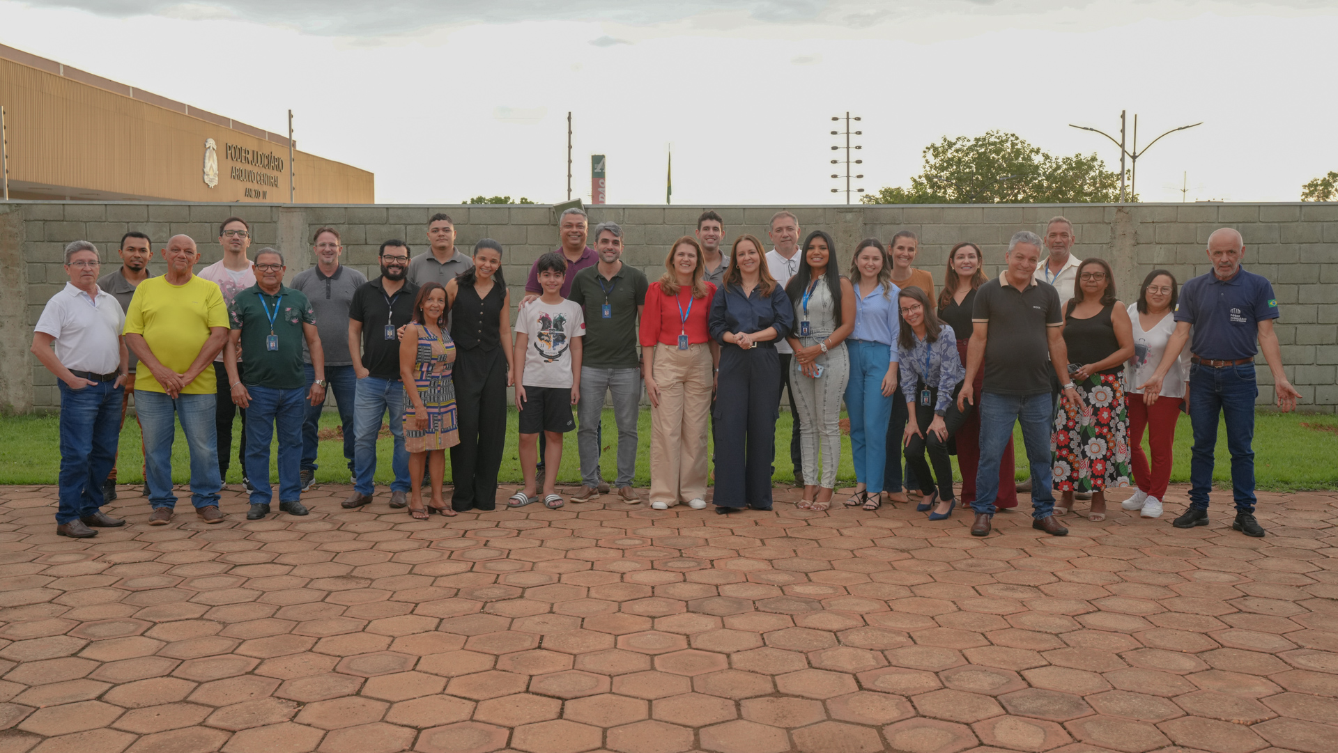 Foto coletiva de um grupo numeroso de pessoas, em sua maioria servidores, reunidas ao ar livre em frente a um prédio do Poder Judiciário. O grupo está alinhado lado a lado, usando roupas casuais e esporte fino, com alguns participantes identificados por crachás. A imagem transmite um clima de integração, união e registro institucional, possivelmente relacionado a uma atividade ou evento coletivo.