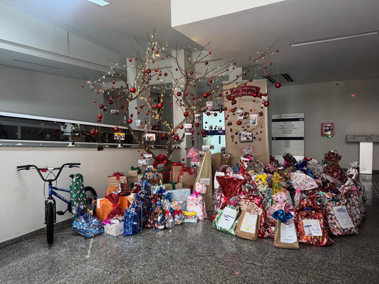 A imagem mostra um espaço interno decorado para o período natalino, com uma árvore estilizada iluminada por luzes e enfeitada com bolas vermelhas. Ao redor, há grande quantidade de presentes embrulhados em papéis coloridos e decorativos, empilhados no chão, muitos deles com cartões e fotos anexadas. O cenário transmite um clima de solidariedade, confraternização e espírito de Natal, marcado pela participação coletiva e pelo cuidado na preparação das doações.