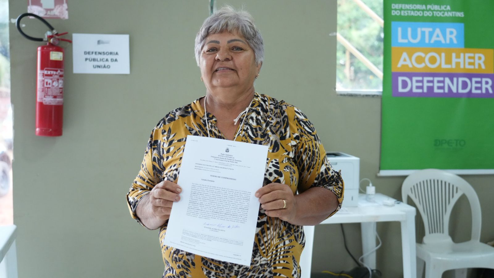 A foto mostra uma mulher segurando um documento à frente do corpo, olhando para a câmera. Ela veste uma blusa estampada em amarelo e preto. Ao fundo, aparecem um extintor, uma placa da Defensoria Pública da União, um banner colorido da Defensoria Pública do Estado do Tocantins e parte de uma mesa com cadeira e equipamentos.