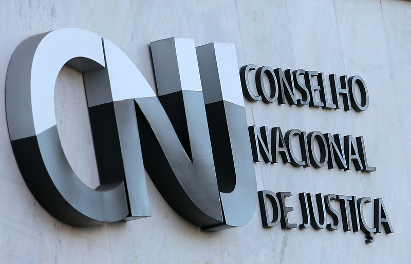 Logotipo do Conselho Nacional de Justiça (CNJ) fixado em uma parede clara. As letras “CNJ” aparecem em destaque, em formato grande, metálico e tridimensional. Ao lado, está escrito “Conselho Nacional de Justiça