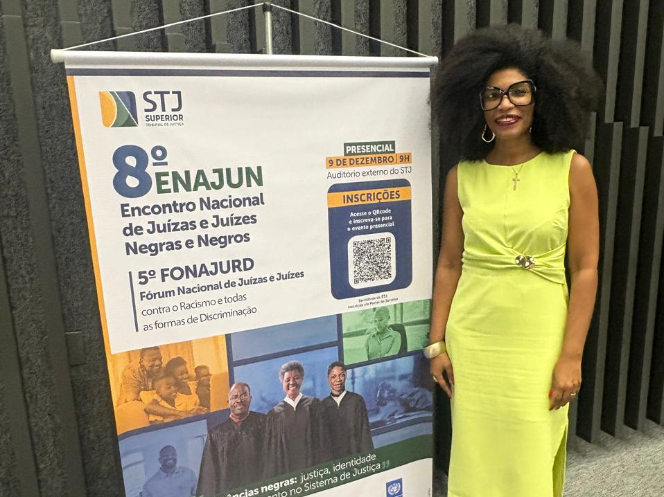 A foto mostra a juíza Renata, uma mulher negra, ao lado de um banner do 8º Enajun e 5º Fonajurd, no STJ. Ela veste um vestido amarelo, usa óculos grandes e tem cabelo black power. O banner traz informações do evento, incluindo data, local e QR Code para inscrições.