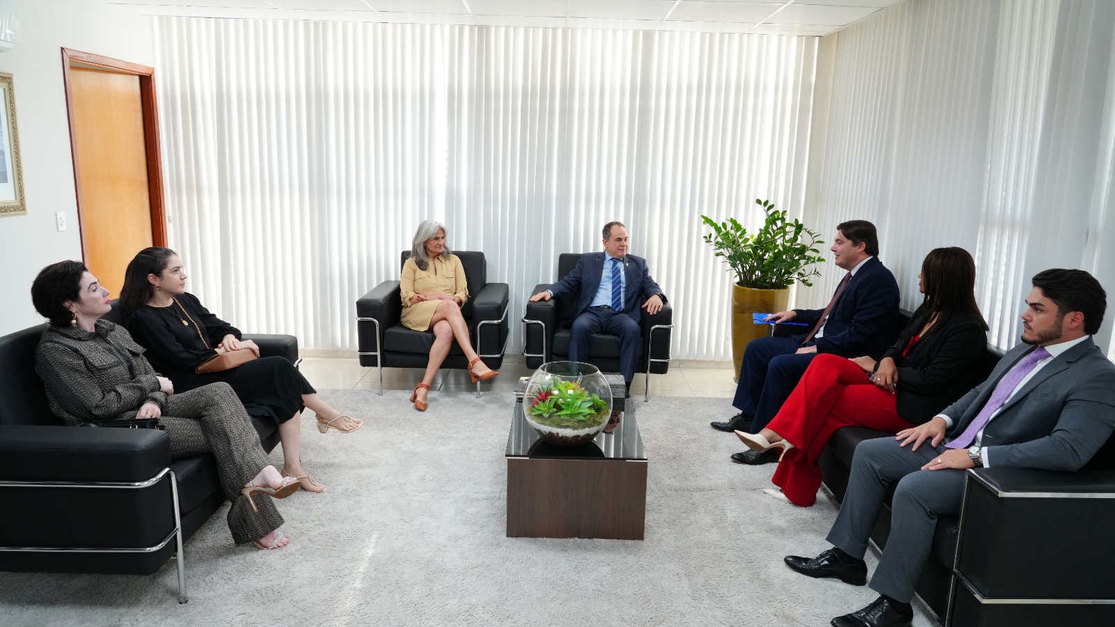 Em uma sala institucional com persianas verticais brancas, sete pessoas estão sentadas em sofás e poltronas pretas, conversando em roda. Ao centro há uma mesa de centro com tampo de vidro e um terrário; ao fundo, um vaso grande com planta verde. Todas usam trajes sociais: à esquerda, duas mulheres com roupas escuras; ao centro, uma mulher de vestido bege e um homem de terno azul; à direita, três pessoas — uma mulher de blazer preto e calça vermelha entre dois homens de terno  acompanham a conversa atentamente.