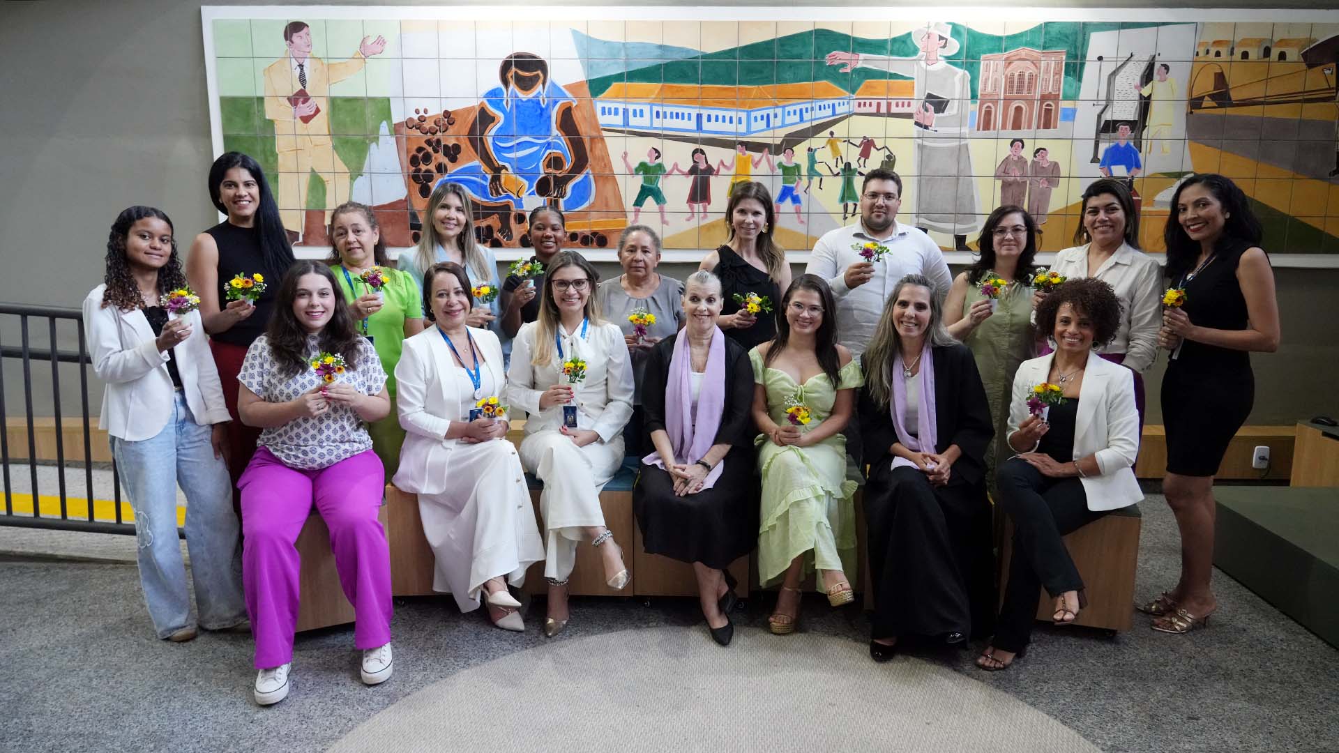 A foto mostra um grupo de mulheres posando em conjunto, sorrindo, em um ambiente interno. Parte delas está sentadas e outra parte em pé, formando duas fileiras. Cada uma segura um pequeno arranjo de flores coloridas. Ao fundo, há um grande painel artístico em azulejos, com ilustrações de cenas culturais e figuras humanas. 
