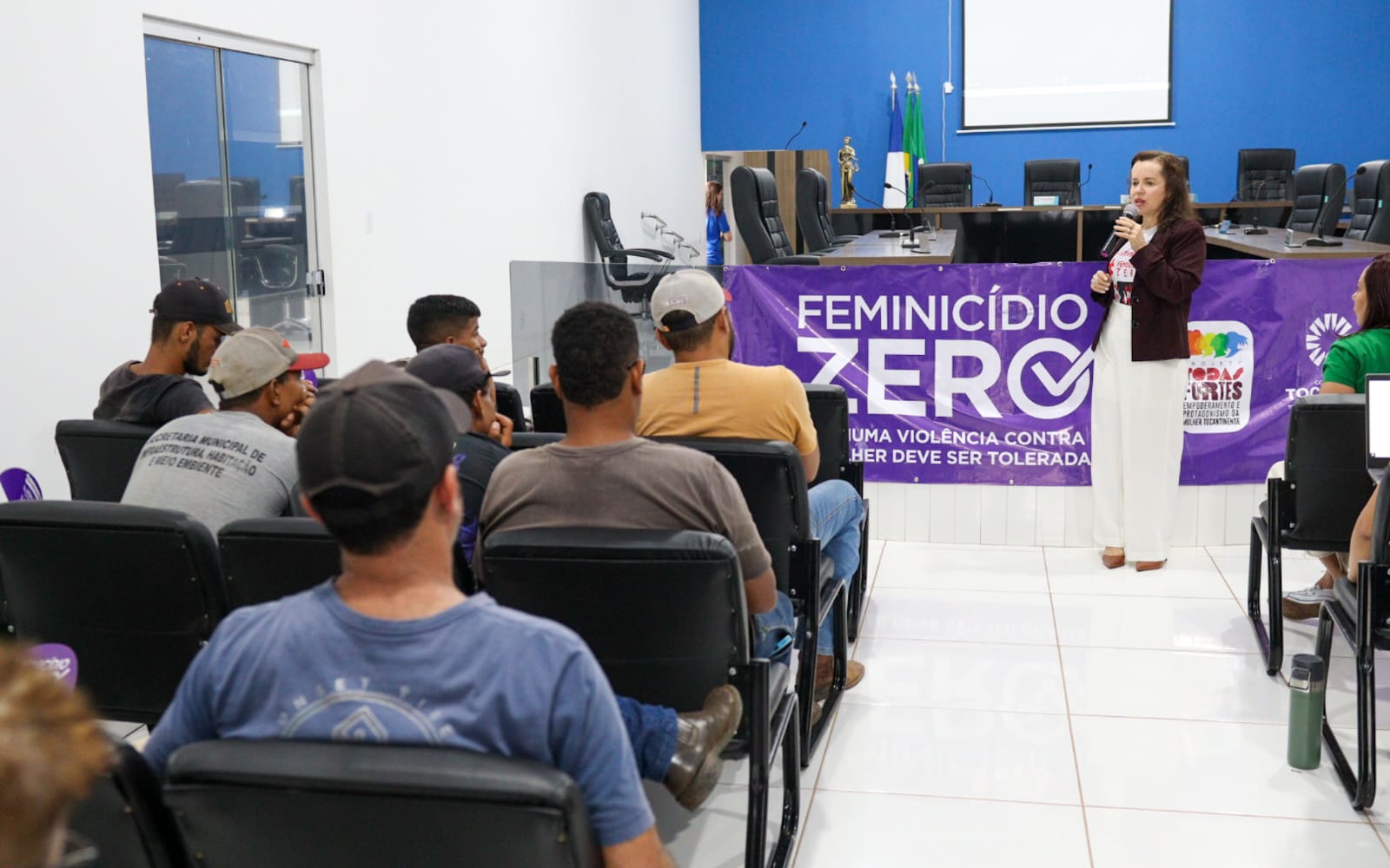 Homens sentados de costas em cadeiras pretas em sala assistindo palestra de mulher de calça branca e blazer preto. Ela está de cabelos soltos, na altura dos ombros e microfone na mão, falando sobre Feminicídio, tema inscrito na faixa roxa atrás dela. Ao fundo, parede azul.