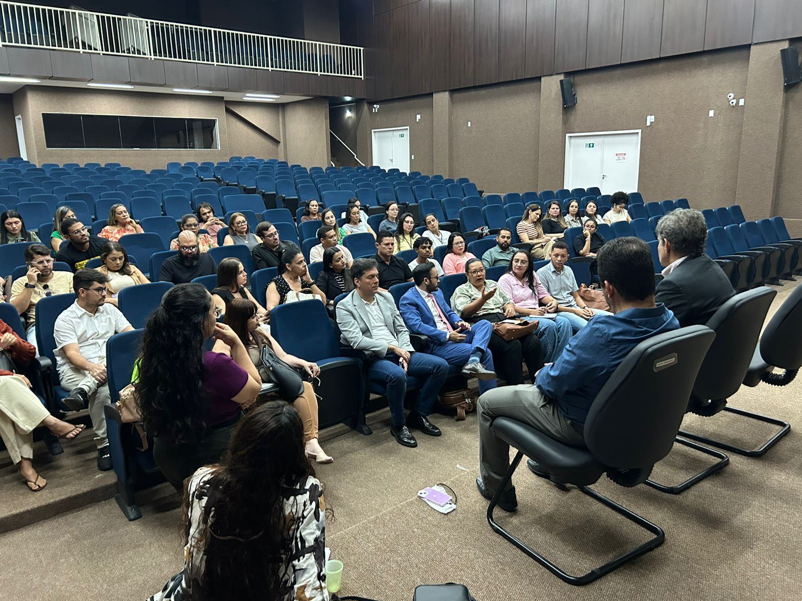 Foto aberta de uma reunião com várias pessoas sentadas em cadeiras azuis, em um auditório