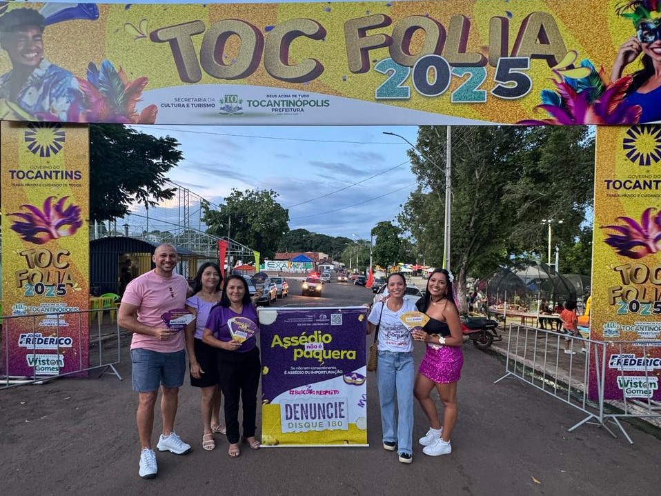 Na imagem um grupo de 5 pessoas em frente a um arco de entrada do evento "TOC FOLIA 2025", do carnaval de Tocantinópolis. Na foto um homem e quatro mulheres estão segurando leques da campanha do TJTO “Assédio não é paquera”, e ao meio tem um banner da mesma campanha.
