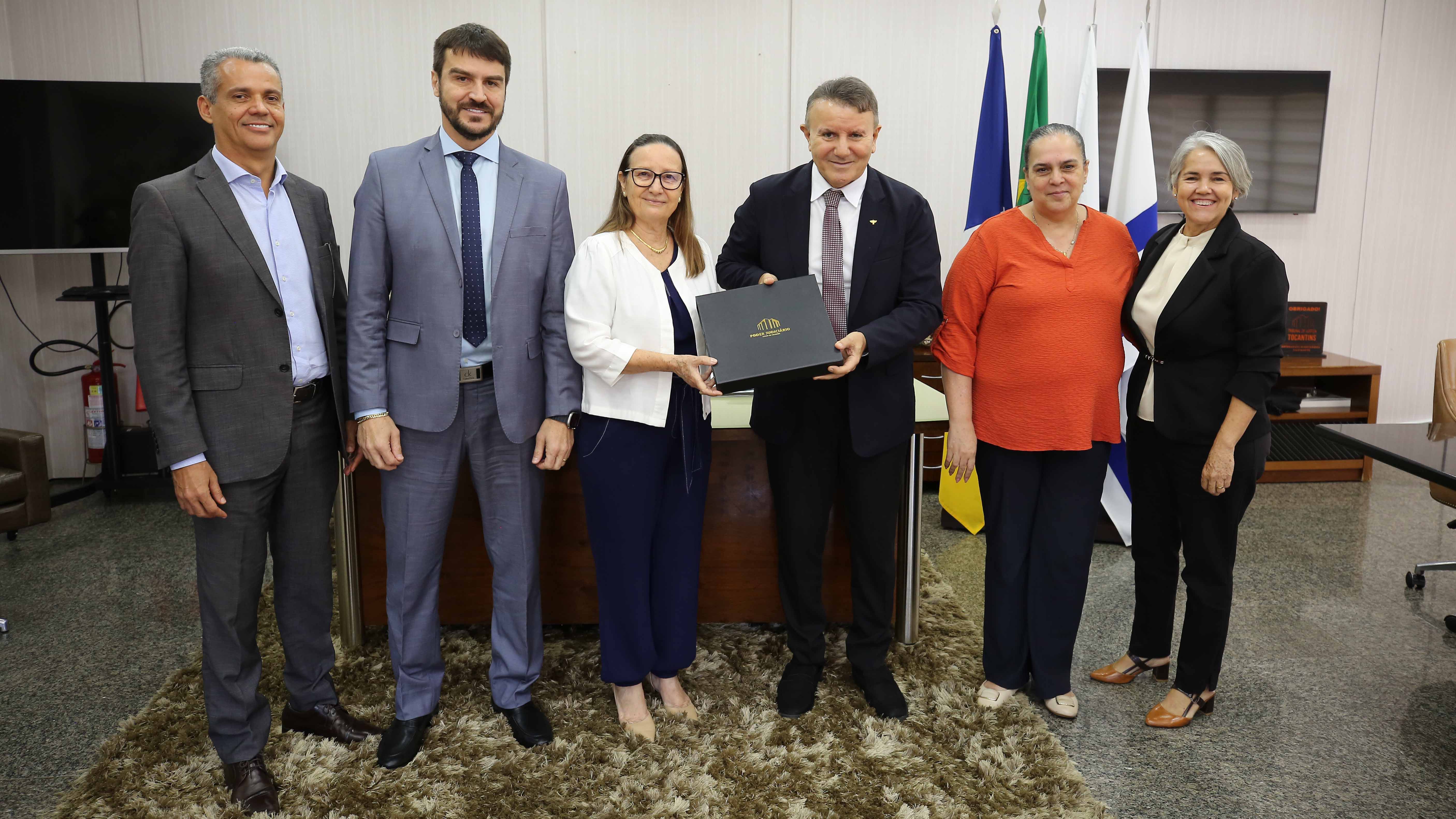 Fotografia colorida onde três mulheres e tr6es homens estão de pé lado a lado posando para uma fotografia enquanto o homem e a mulher do meio do grupo seguram uma caixa retangular preta. Eles estão em um sala com bandeiras e uma mesa ao fundo, ao lado de uma televisão. No chão, um carpete de cor amarelo escuro 