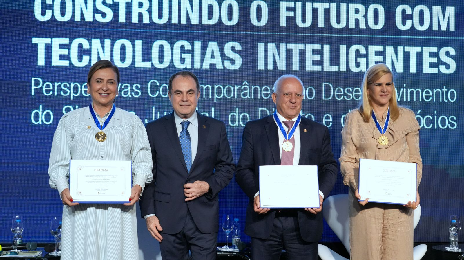 Fotografia colorida onde dois homens e duas mulheres vestidos em roupas sociais estão em frente a um painel azul escuro repleto de palavras. Duas das mulheres e um dos homens estão segurando certificados e com medalhas no pescoço 