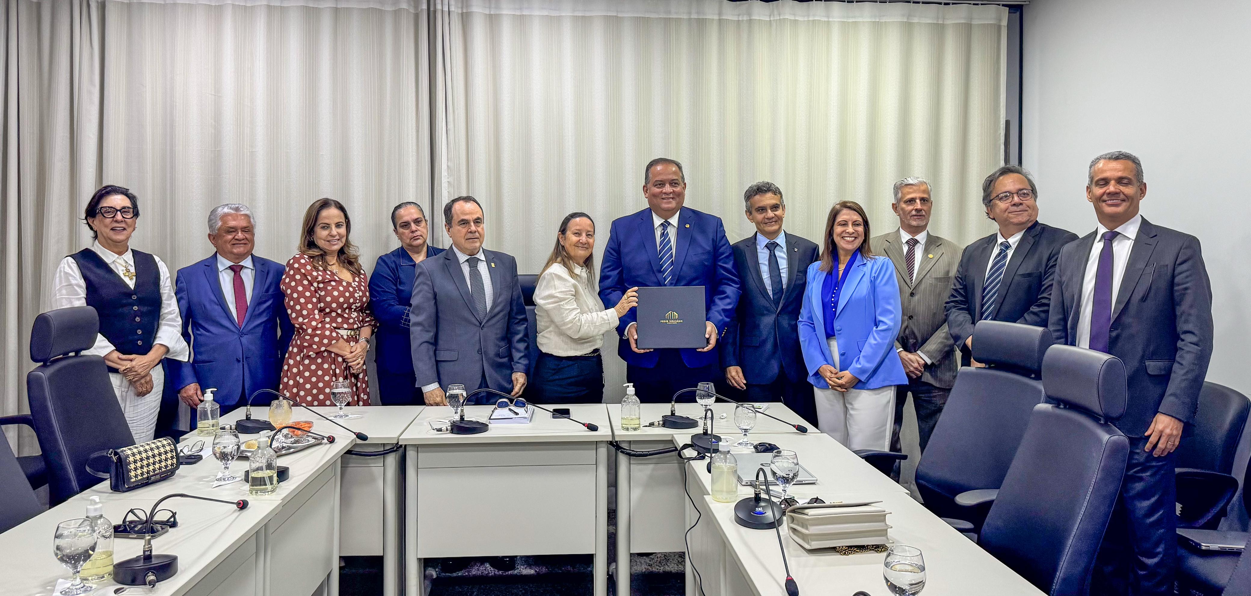Foto aberta em que aparecem 12 pessoas, entre homens e mulheres, posando para foto. Eles estão em uma sala com mesas, cadeiras e microfones sobre as mesas. Ao centro, um deles segura um objeto entregue por uma mulher que está ao seu lado. Todos usam roupas formais.