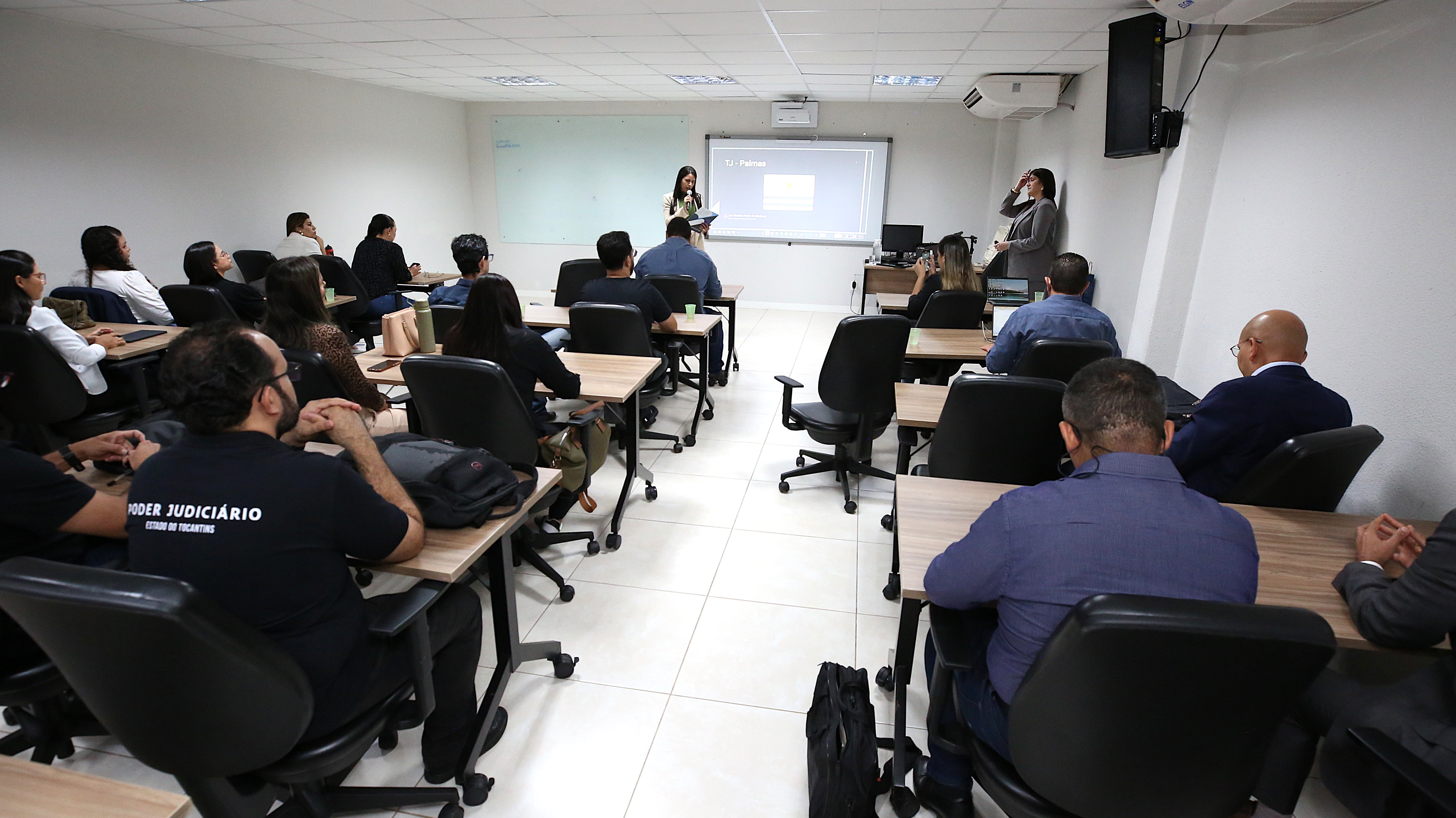 Fotografia colorida onde um grande grupo de pessoas estão sentados em uma sala branca observando uma mulher de roupa social cinza que fala a frente
