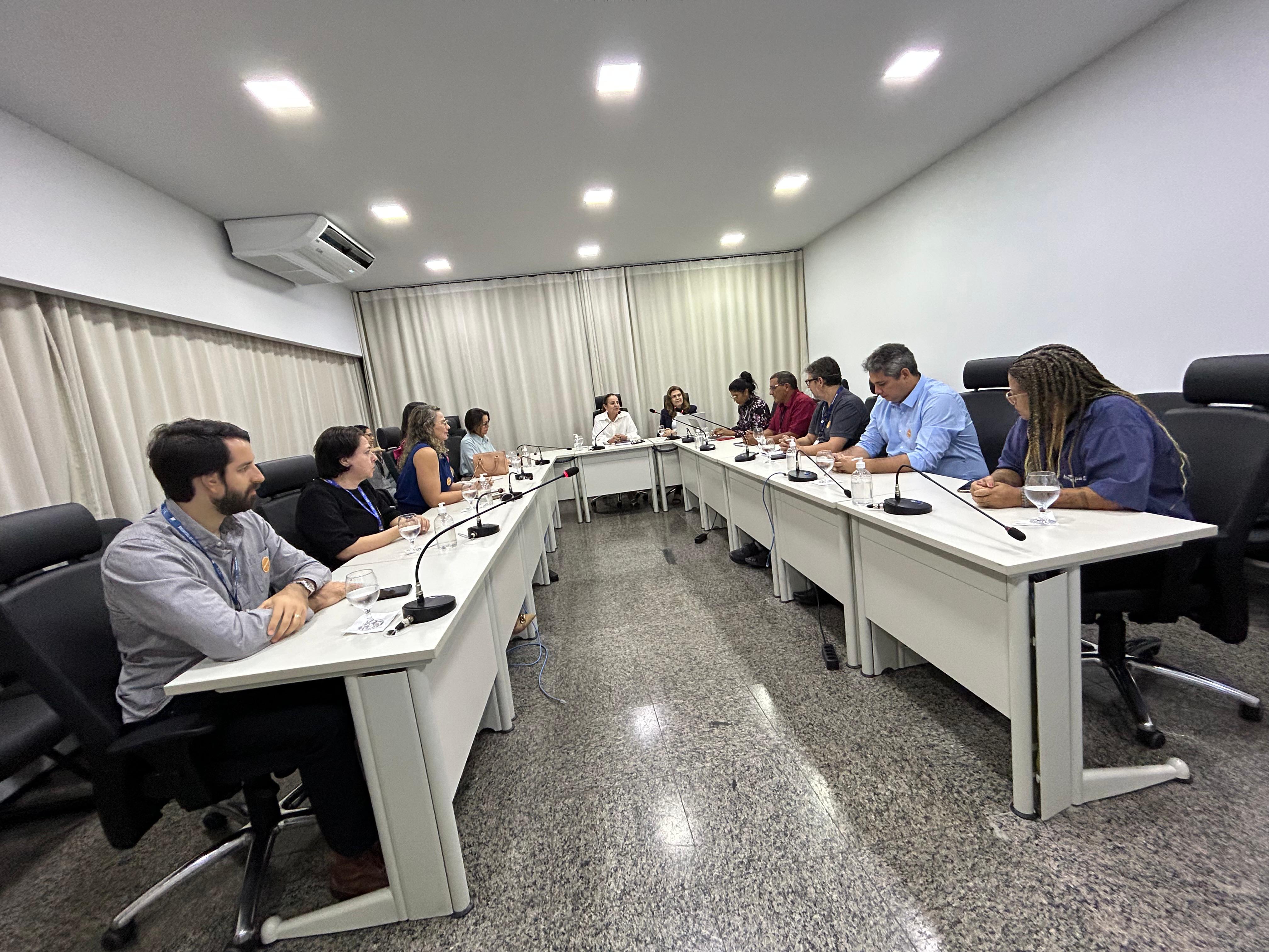 Fotografia colorida de uma sala branca com uma cortina de fundo em cor de creme. Ao centro, uma mesa em formato de U está disposta com várias pessoas sentadas em volta dela em conversa.