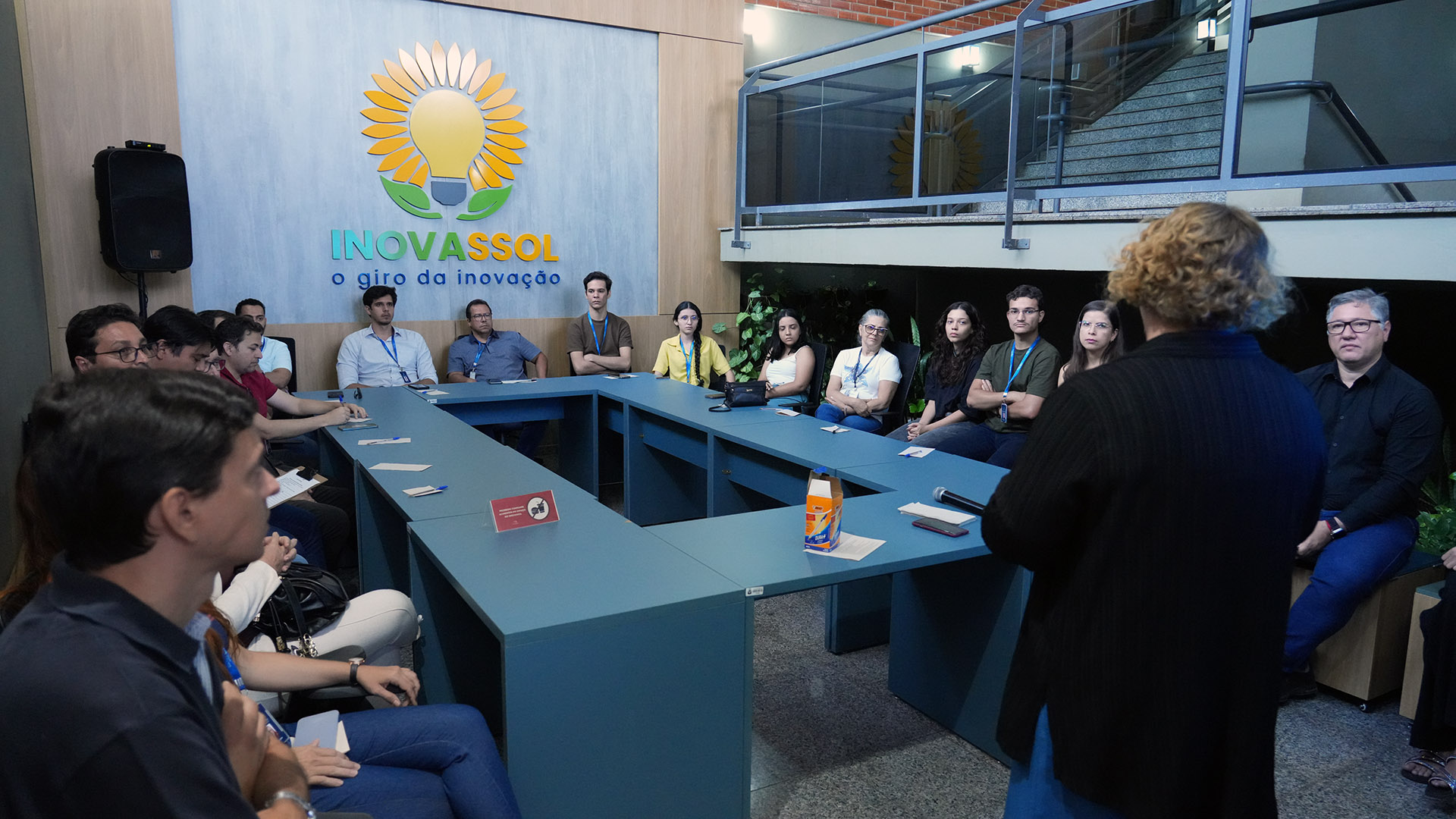 Imagem de uma reunião no espaço Inovassol, com participantes sentados em formato de “U”, atentos à fala de uma pessoa em pé. Ao fundo, a logomarca do Inovassol com o slogan “o giro da inovação”. Ambiente moderno, com plantas e escada lateral. Clima de concentração.