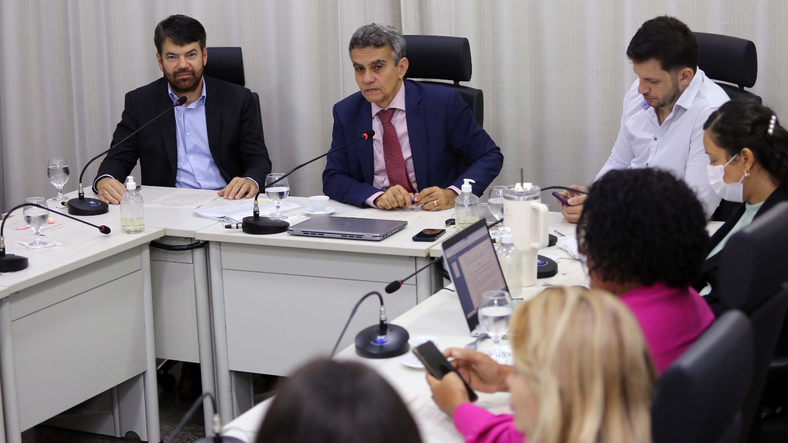 Uma reunião formal acontece em uma sala com cortinas brancas ao fundo. Duas pessoas estão em destaque na mesa central: um homem de terno azul e gravata vermelha, que fala ao microfone, e outro homem de blazer preto e camisa azul, que escuta atentamente. Ao redor da mesa, há mais participantes, incluindo uma mulher de máscara branca e outra de blusa rosa. A mesa está equipada com microfones, notebooks, garrafas de água, e frascos de álcool em gel.