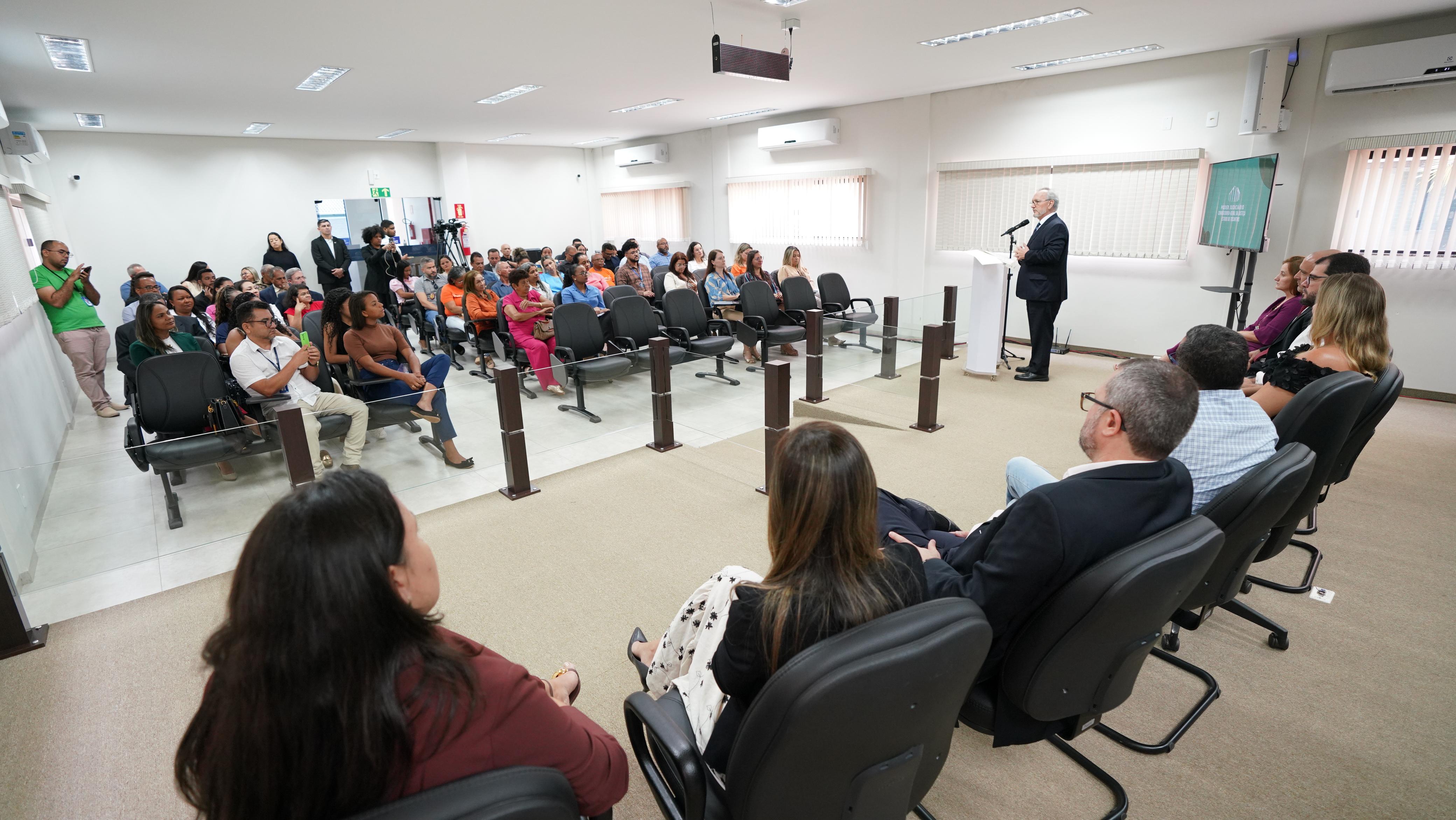 Fotografia colorida onde um grupo de pessoas estão sentadas em cadeiras pretas em um auditório. Alguns homens e mulheres estão sentados na frente do grupo em cadeiras pretas em cima de um palco enquanto um homem vestido de terno completo, com barba e cabelos grisalhos fala atrás de um púlpito 