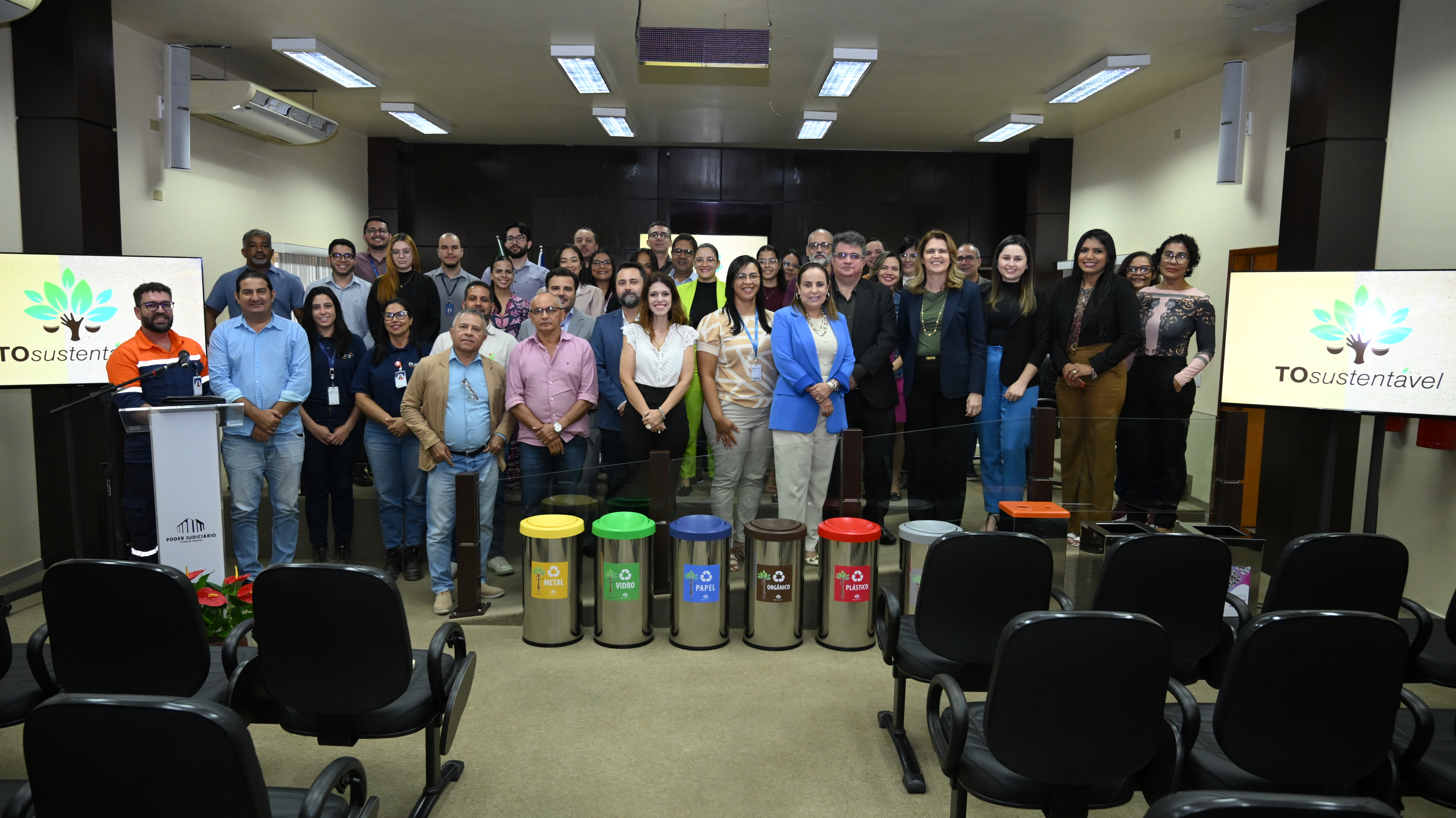 Fotografia colorida onde um grande grupo de pessoas se reúnem para tirar uma foto em frente a lixeiras sustentáveis. As pessoas estão dentro de uma sala de reuniões com cadeiras pretas em sua frente. O teto e as paredes são amarelas claro com luzes e detalhes em madeira escuro 
