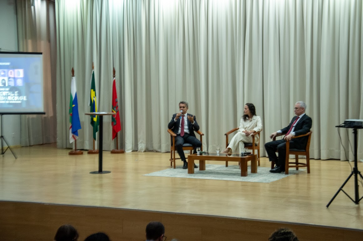 A imagem mostra um palco com três pessoas, dois homens e uma mulher ao centro, sentadas em cadeiras, em frente a uma plateia. Há uma pequena mesa baixa entre as duas pessoas do centro e da direita. No fundo do palco, há uma tela, onde estão sendo exibidas imagens ou informações. Há bandeiras de diferentes países no palco. As cortinas brancas cobrem as paredes do fundo do palco.
