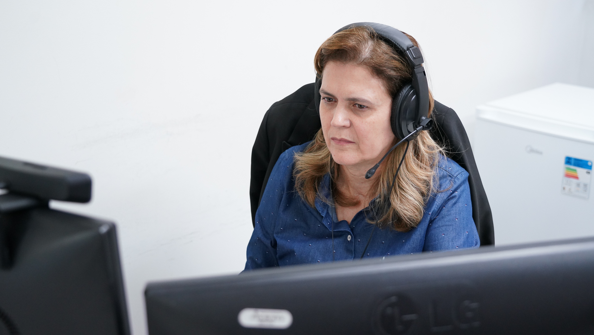 Uma mulher com fones de ouvido, sentada em frente a um computador, está concentrada, apresenta uma expressão séria e atenta, e parece estar imersa em alguma tarefa em seu computador. Usa uma camisa de manga comprida de cor azul-acinzentada e seu cabelo é castanho claro e levemente ondulado. Seu rosto é o ponto focal da imagem e parece em calma e concentração.
